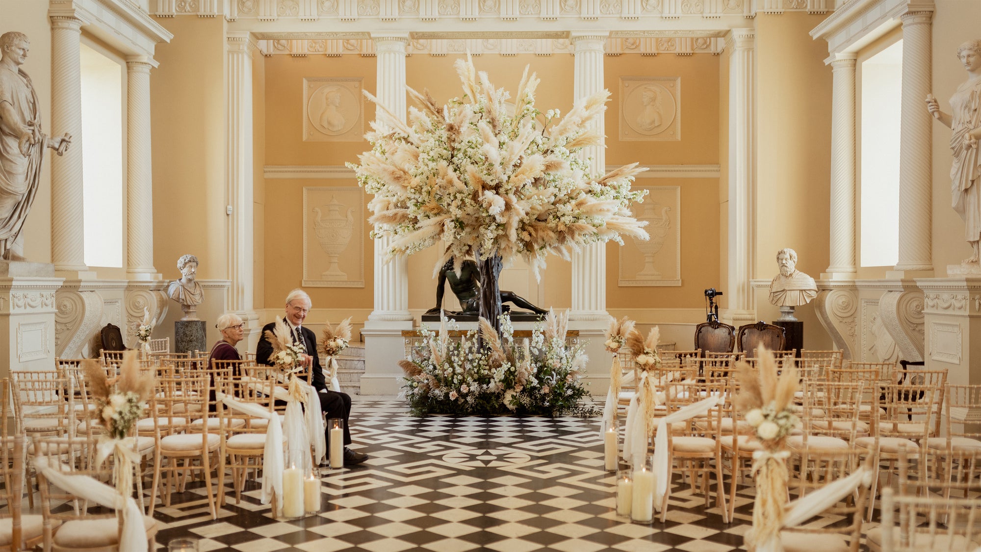 Wedding ceremony setup at Syon Park, London, with dramatic pampas grass centerpiece, gold chiavari chairs, and neoclassical sculpture gallery - Floral Design by Amaranté London