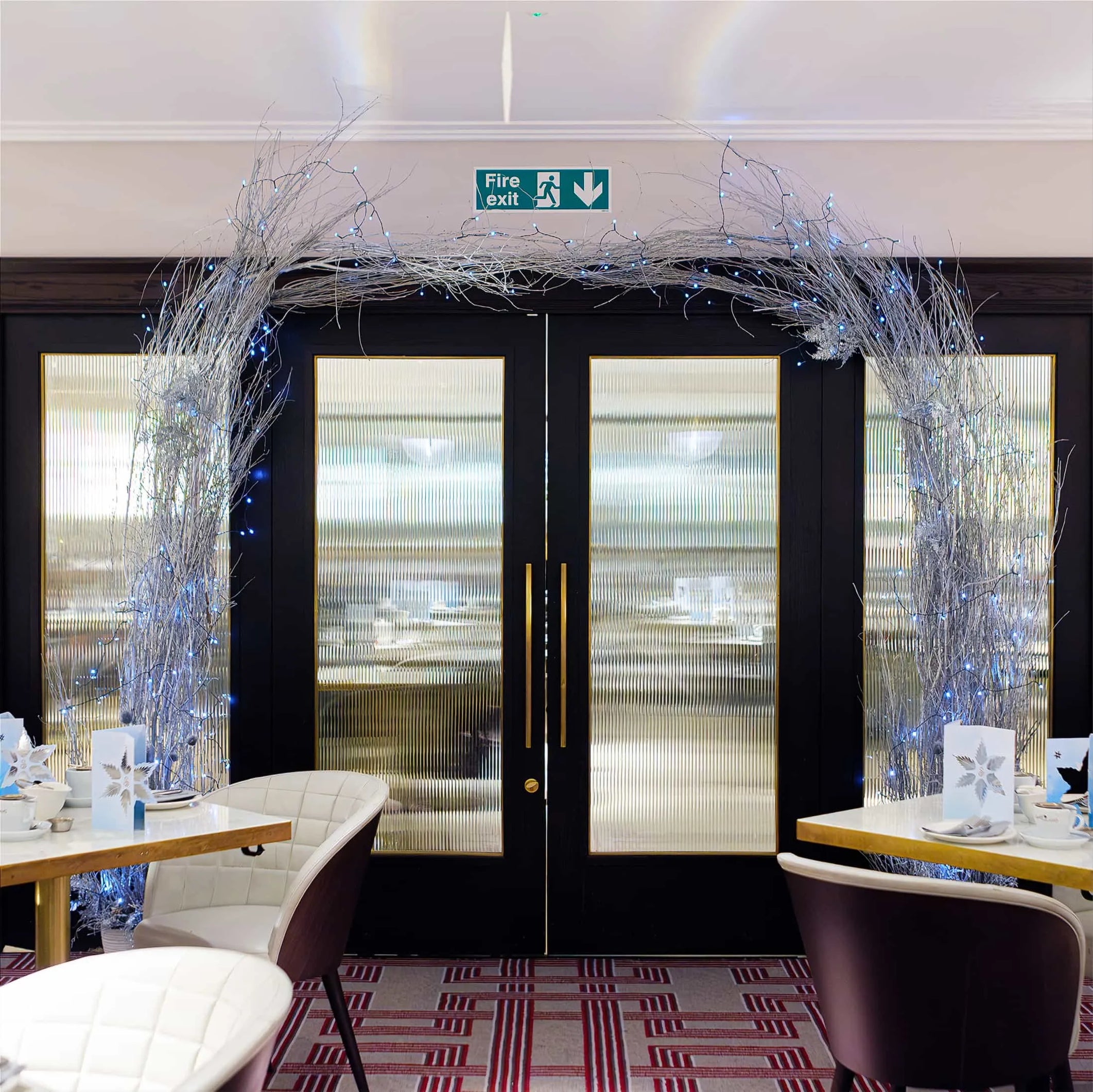 An elegant bespoke floral arch created by Event Florist Amaranté London frames the entrance to a dining area in the Strand Palace Hotel, featuring a lattice of silver branches sprinkled with twinkling lights, adding a festive and sophisticated atmosphere. The arch, set against the contrast of the rich maroon walls and the dark door, invites guests into the space, promising a cheerful and relaxing dining experience during the Christmas holidays.