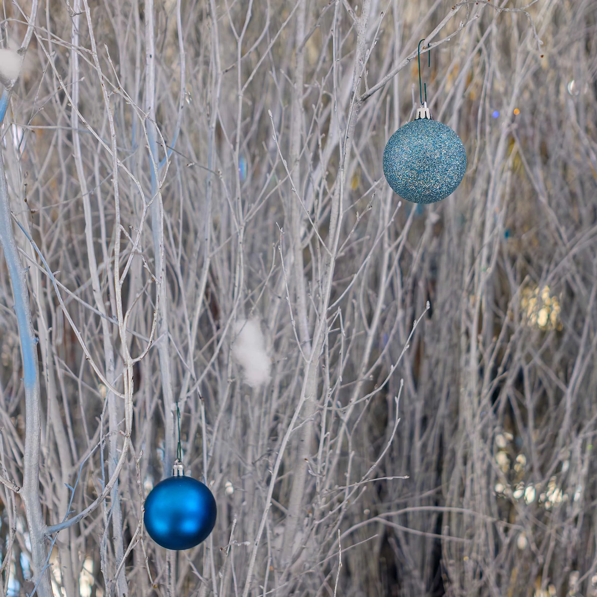 Image of a close-up of a bespoke floral arrangement featuring natural birch branches painted in a frosty silver, set against a soft-focus background, with two Christmas baubles, one in deep sapphire blue and the other glittering with a dusting of snow-like sparkles, that hang amidst the branches. This Christmas Floral installation embodies Christmas's serene and crisp essence as envisioned for the Strand Palace Hotel's holiday decor, designed, produced and installed by Amaranté London -Event Florist.
