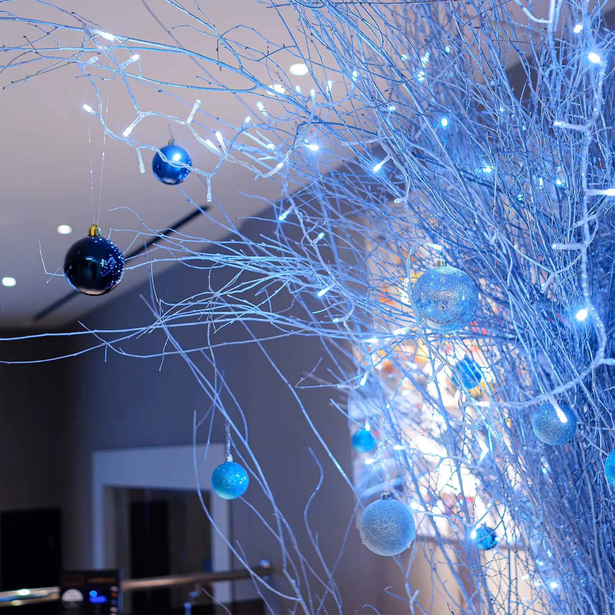 A stunning blue and silver Christmas floral arrangement at the Strand Palace Hotel in London featuring blue baubles and fairy lights intertwined with white birch branches, creating a winter wonderland atmosphere. Bespoke event floristry by Amaranté London.