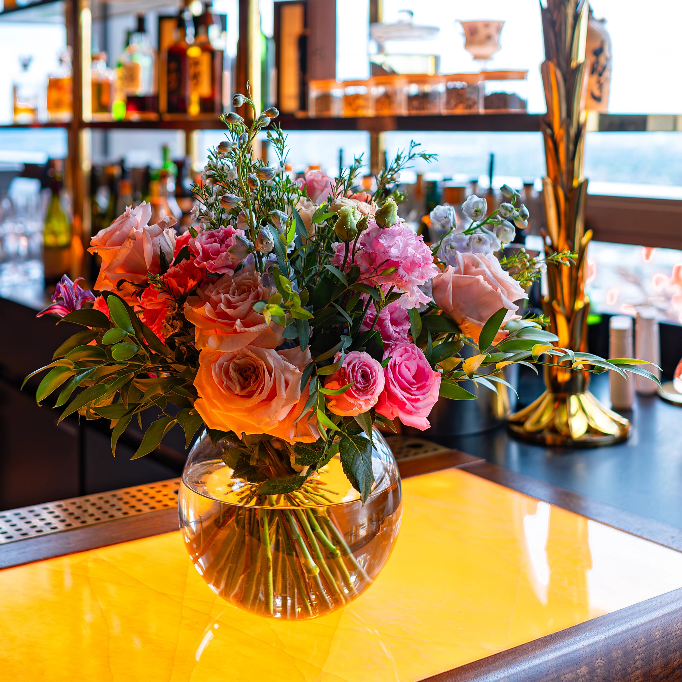 Luxurious flower arrangement on a bar counter at Sushi Samba, featuring a vibrant mix of pink roses, peach blooms, and lush greenery, perfectly complementing the sophisticated interior décor of the venue for a birthday event celebration - Designed, created and installed by Event Florist Amaranté London.