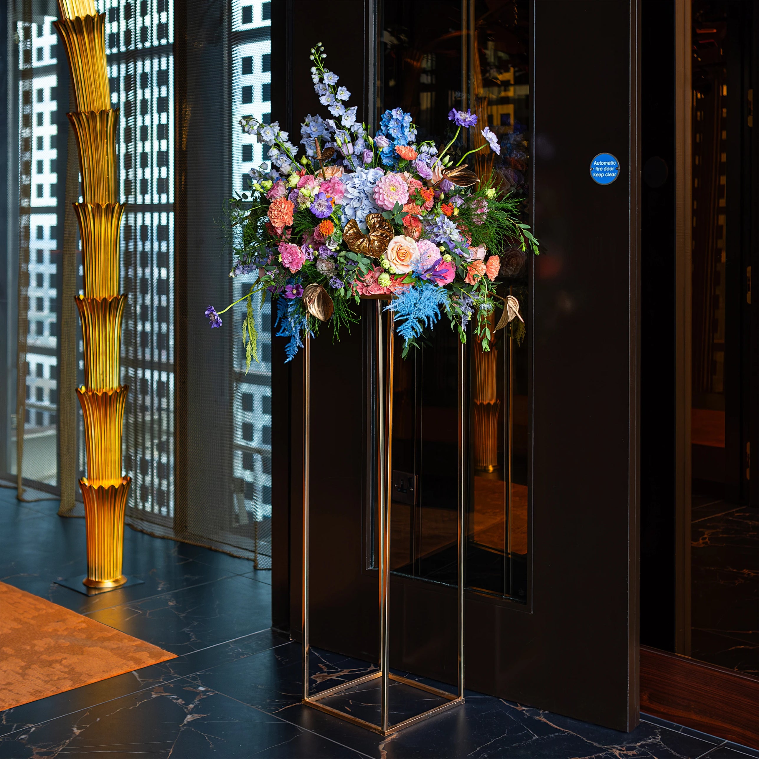 A stunning large floral arrangement at the entrance of Sushi Samba for an exclusive birthday celebration, featuring a mix of blue delphiniums, pink roses, and golden anthurium leaves, creates an impressive and vibrant welcoming display - Amaranté London
