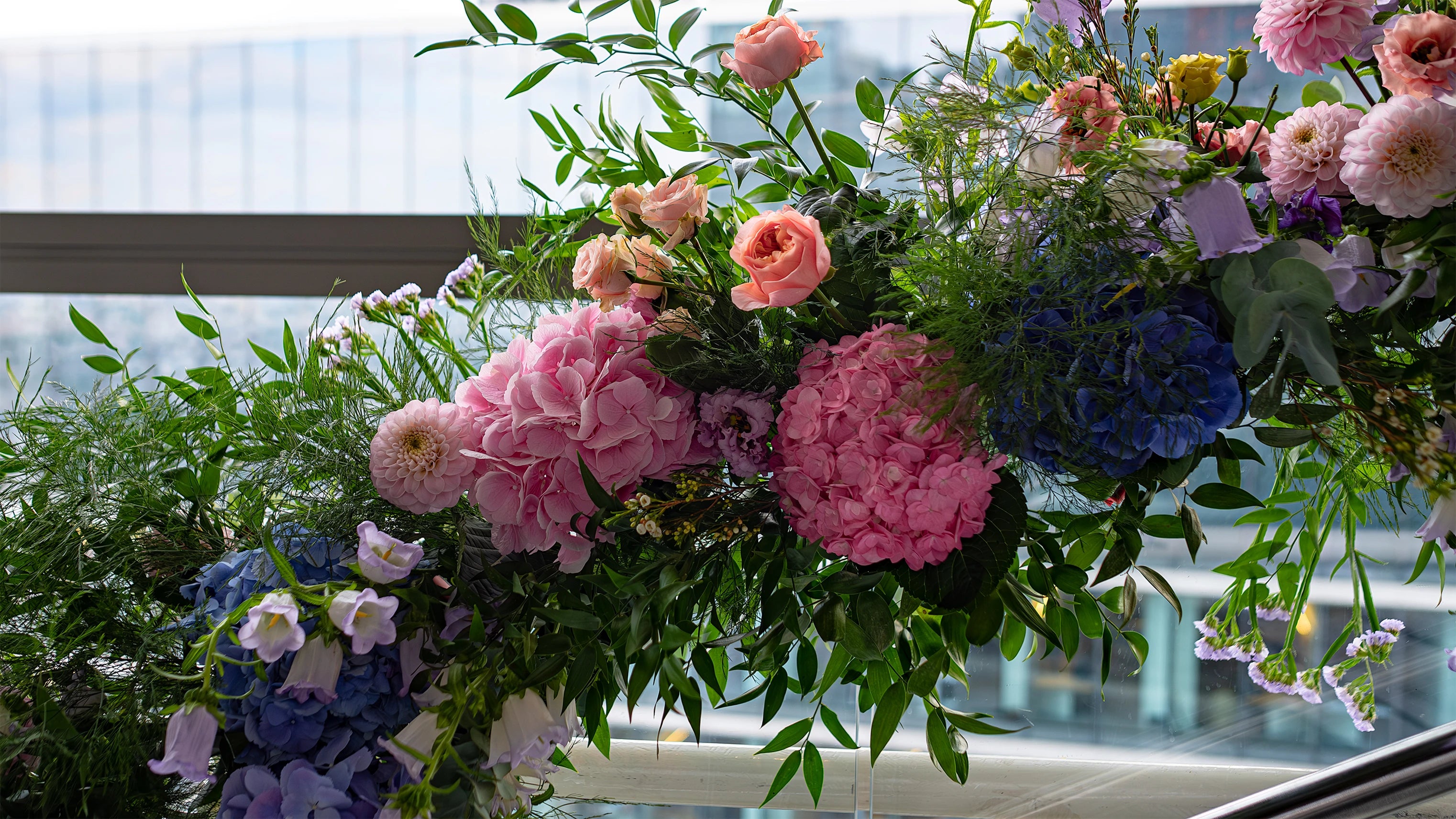 A lush floral arch installation by Amarante London at Sushi Samba for a birthday celebration features pink and blue hydrangeas, peach roses, and green foliage, beautifully framing a window of the venue.