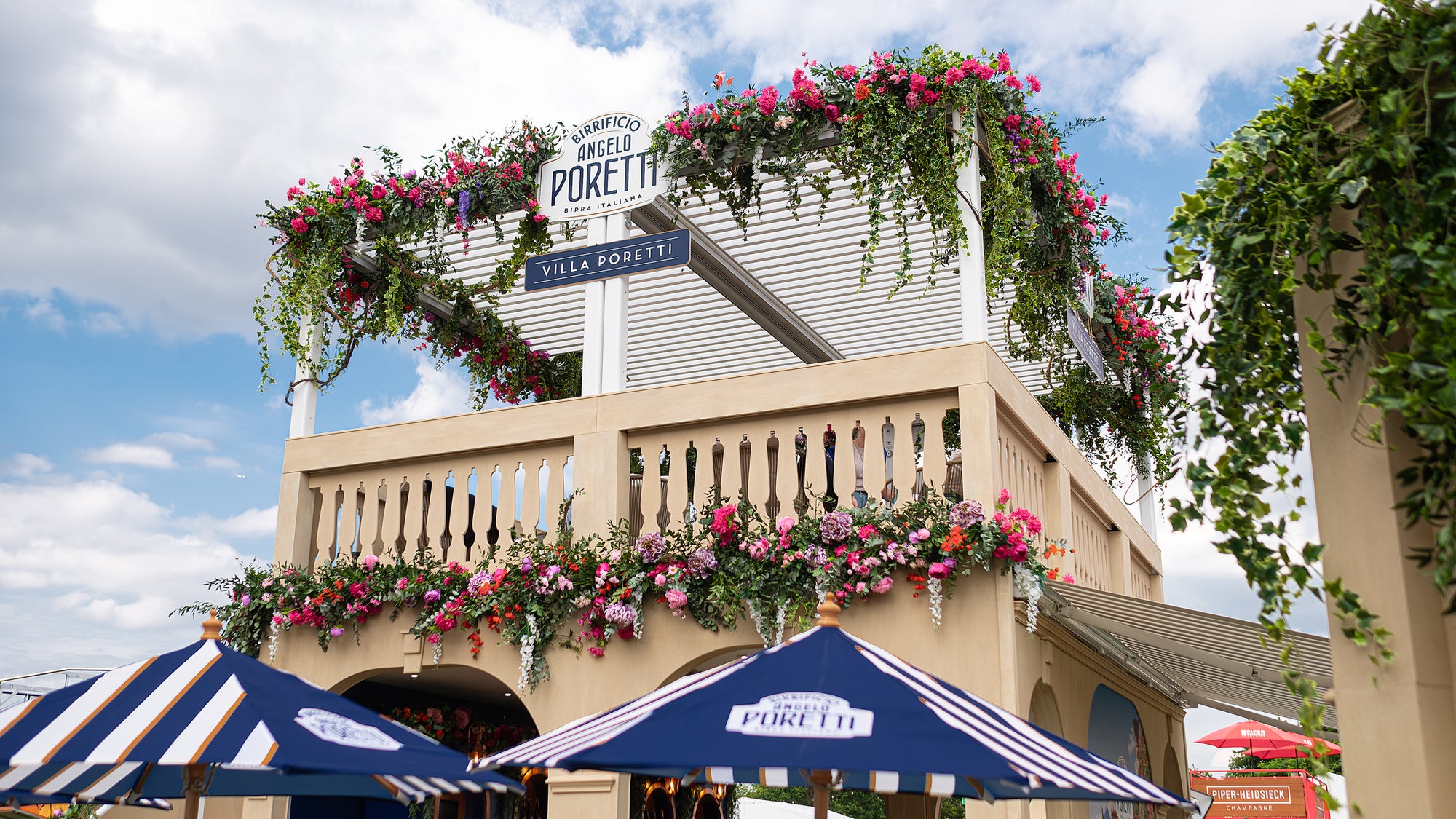 The Upper terrace of the Angelo Poretti stand decorated with pink, red, and orange flowers and green ivy by event florist Amaranté London. The floral decor enhanced the Mediterranean feel and created an inviting atmosphere at Regent’s Park Taste Festival, a fine example of event floristry.