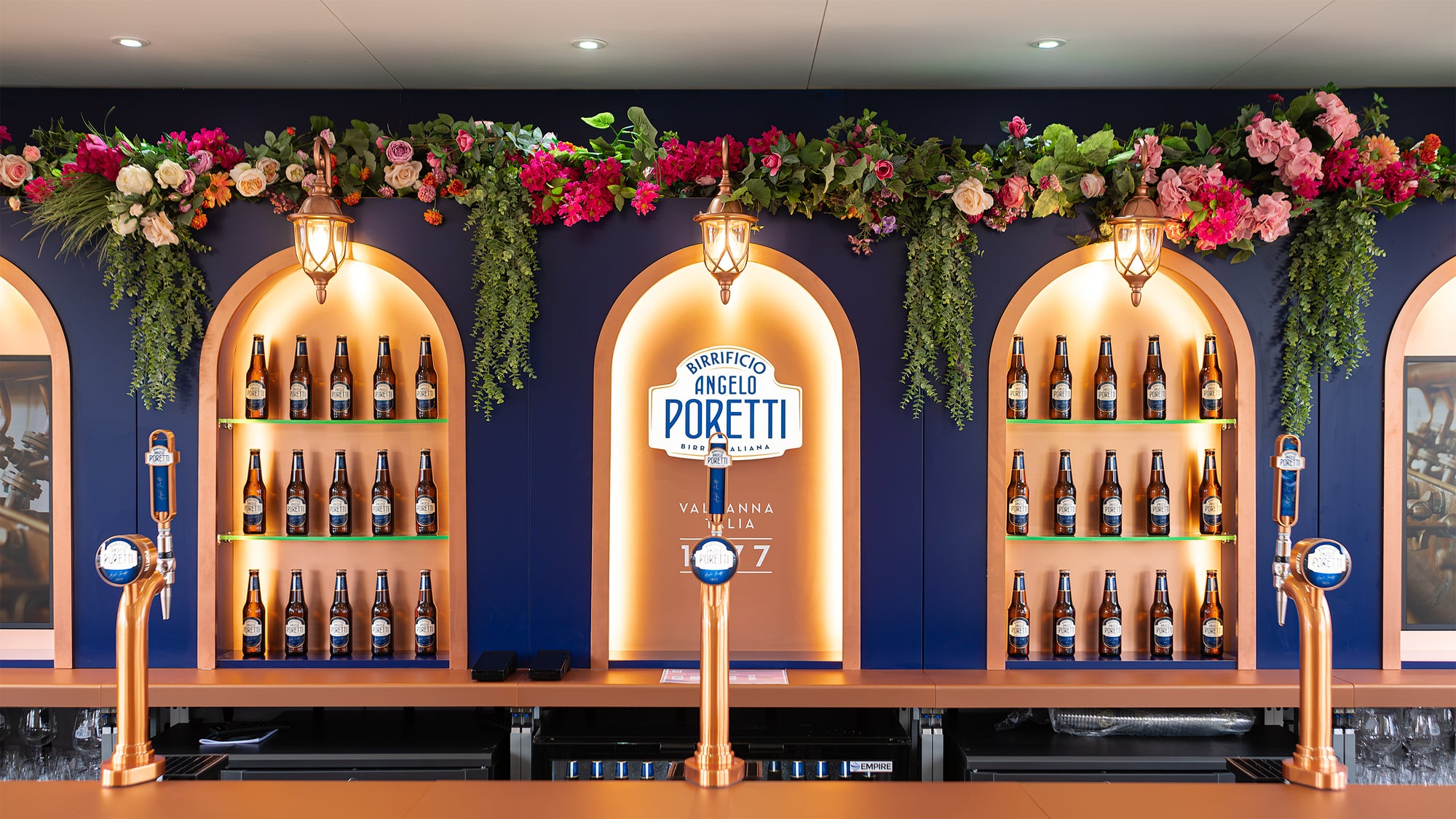 Angelo Poretti beer stand at Regent’s Park Taste Festival decorated by Event Florist Amaranté London. The arches are adorned with vibrant pink, red, and orange flowers and lush green foliage. Blue awnings and wicker chairs enhance the cheerful and welcoming Mediterranean ambiance. Event floristry by Amaranté London.