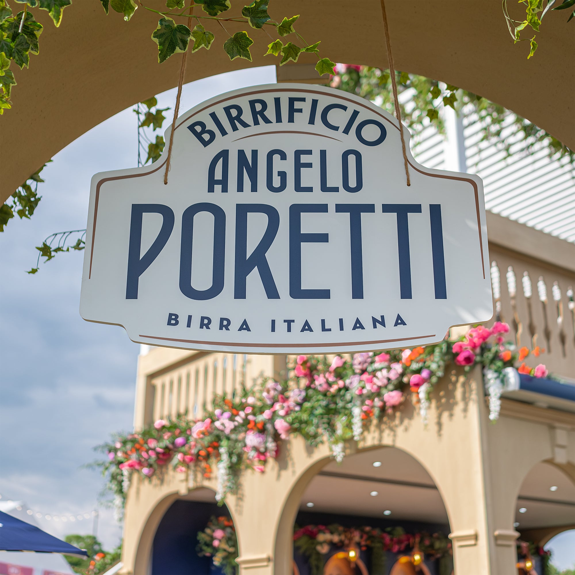 Close-up of Angelo Poretti sign at Regent’s Park Taste Festival Event, framed by lush ivy and vibrant floral arrangements in pink and orange by Florist for events Amaranté London. The floral decor beautifully complements the stand’s Mediterranean design, showcasing Amaranté London’s expertise.