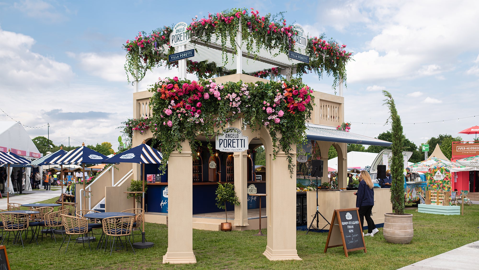 Angelo Poretti beer stand at Regent’s Park Taste Festival decorated by Event Florist Amaranté London. The archway is adorned with vibrant pink, red, and orange flowers and lush green foliage. Blue awnings and wicker chairs enhance the Mediterranean ambiance. An exquisite example of Amaranté London’s event floristry.