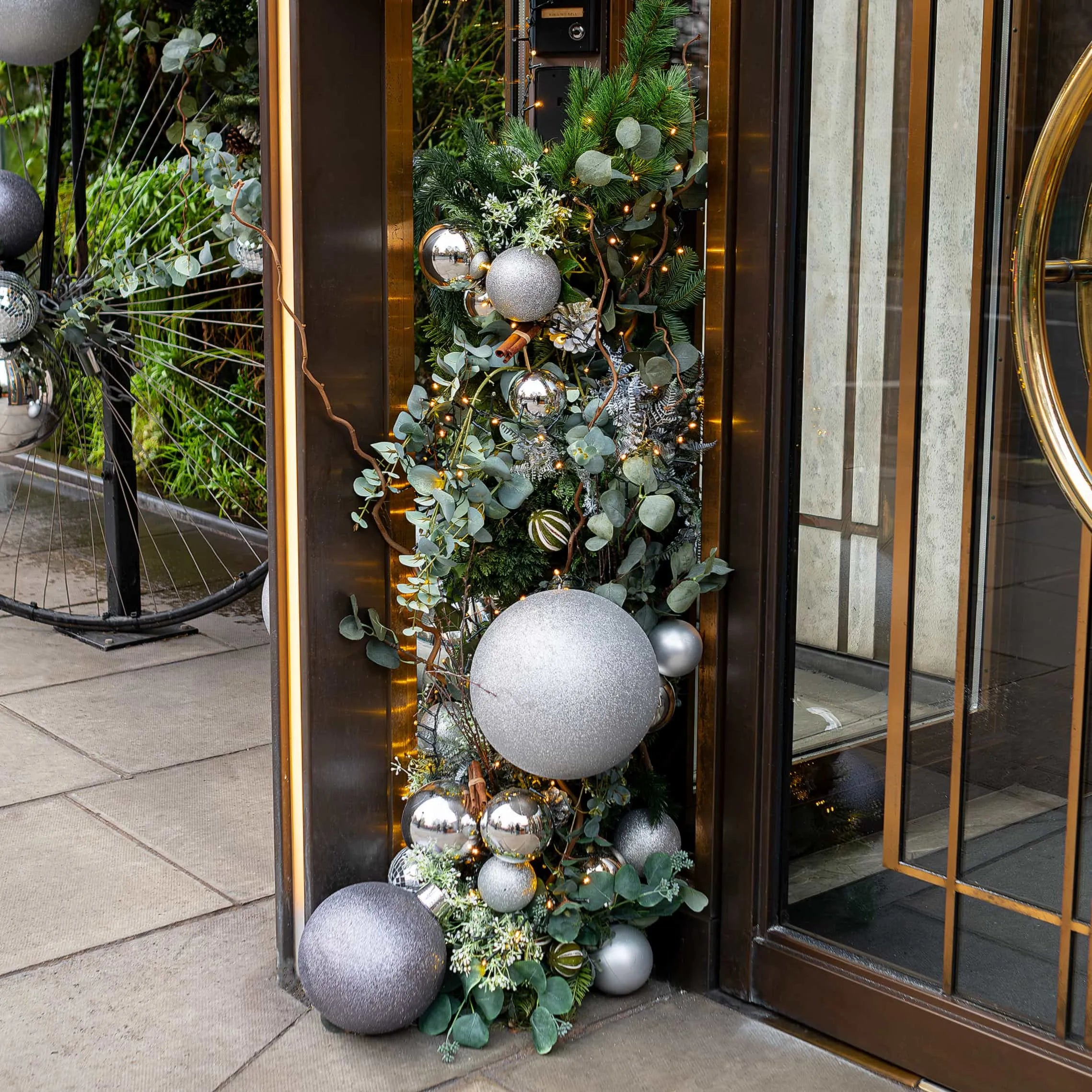 A detailed view of 'The Athenaeum', 5 star hotel in London, entrance adorned with a bespoke festive floral installation designed and installed by event florist Amaranté London. This bespoke floral arrangement showcases a cascade of large silver baubles nestled among eucalyptus leaves and pine foliage, with fairy lights woven throughout to create a captivating and luxurious welcome during the Christmas Holidays.