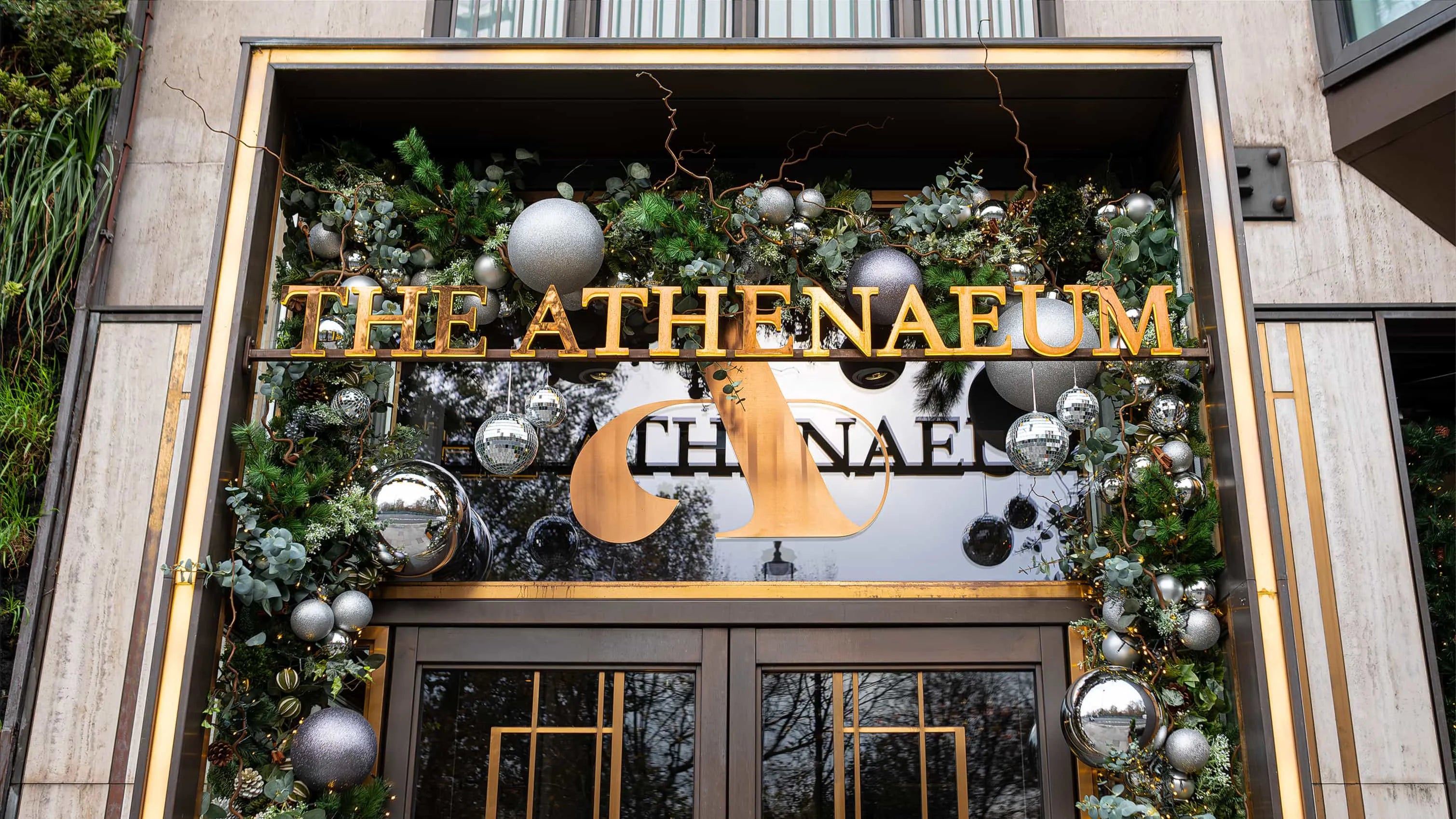 The entrance of 'The Athenaeum' hotel adorned with bespoke floral arrangements designed, produced, and installed by event florist Amaranté London, featuring a festive display of silver ornaments, twinkling lights, and a lush garland of greenery and eucalyptus leaves, highlighting the hotel's commitment to luxury and exclusivity.