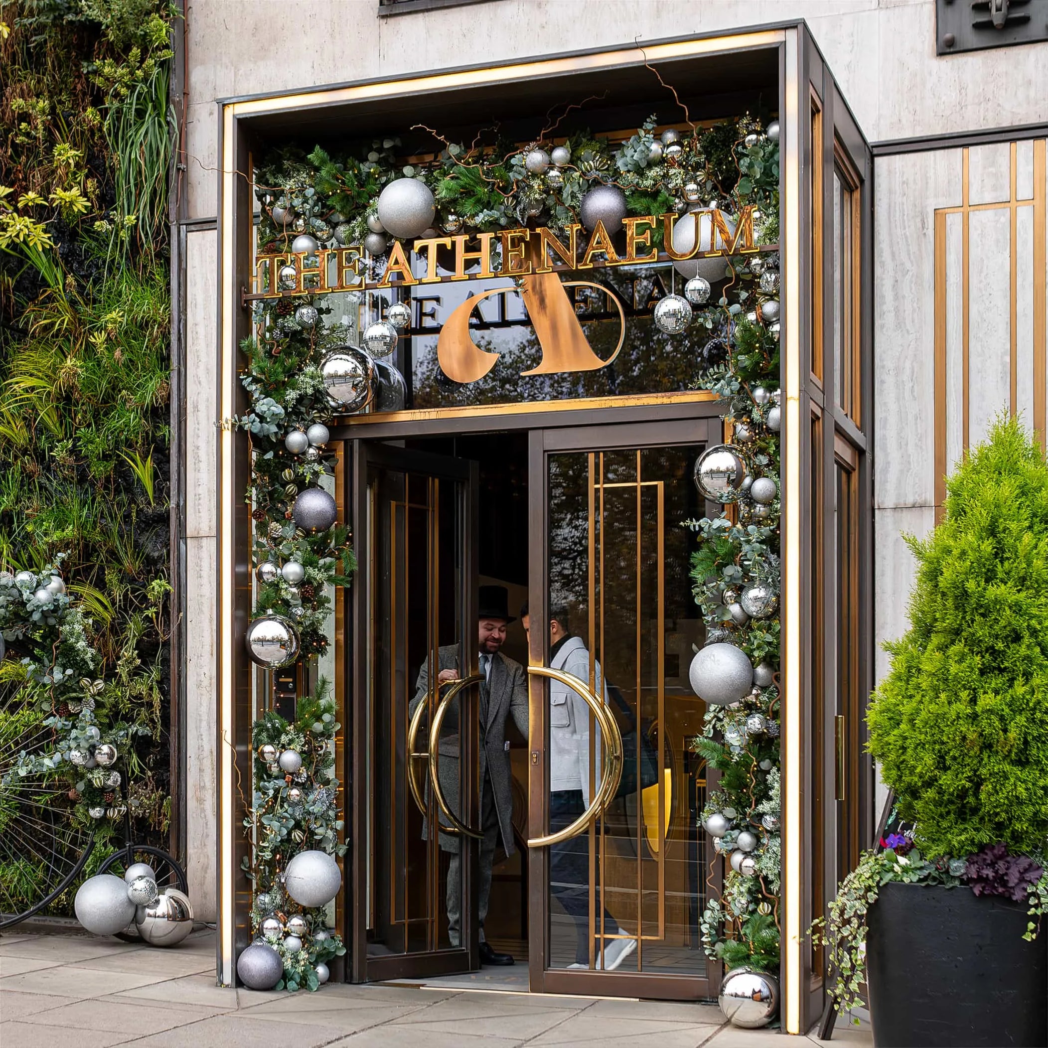 The grand entrance of 'The Athenaeum' hotel, framed by Amaranté London's bespoke holiday floral design, boasts an archway of verdant greenery, interlaced with an array of matte and shiny silver baubles. The festive arrangement creates a luxurious and welcoming atmosphere, complemented by the hotel's signature golden signage and the natural elegance of a vertical living wall, as a guest steps through the revolving doors.