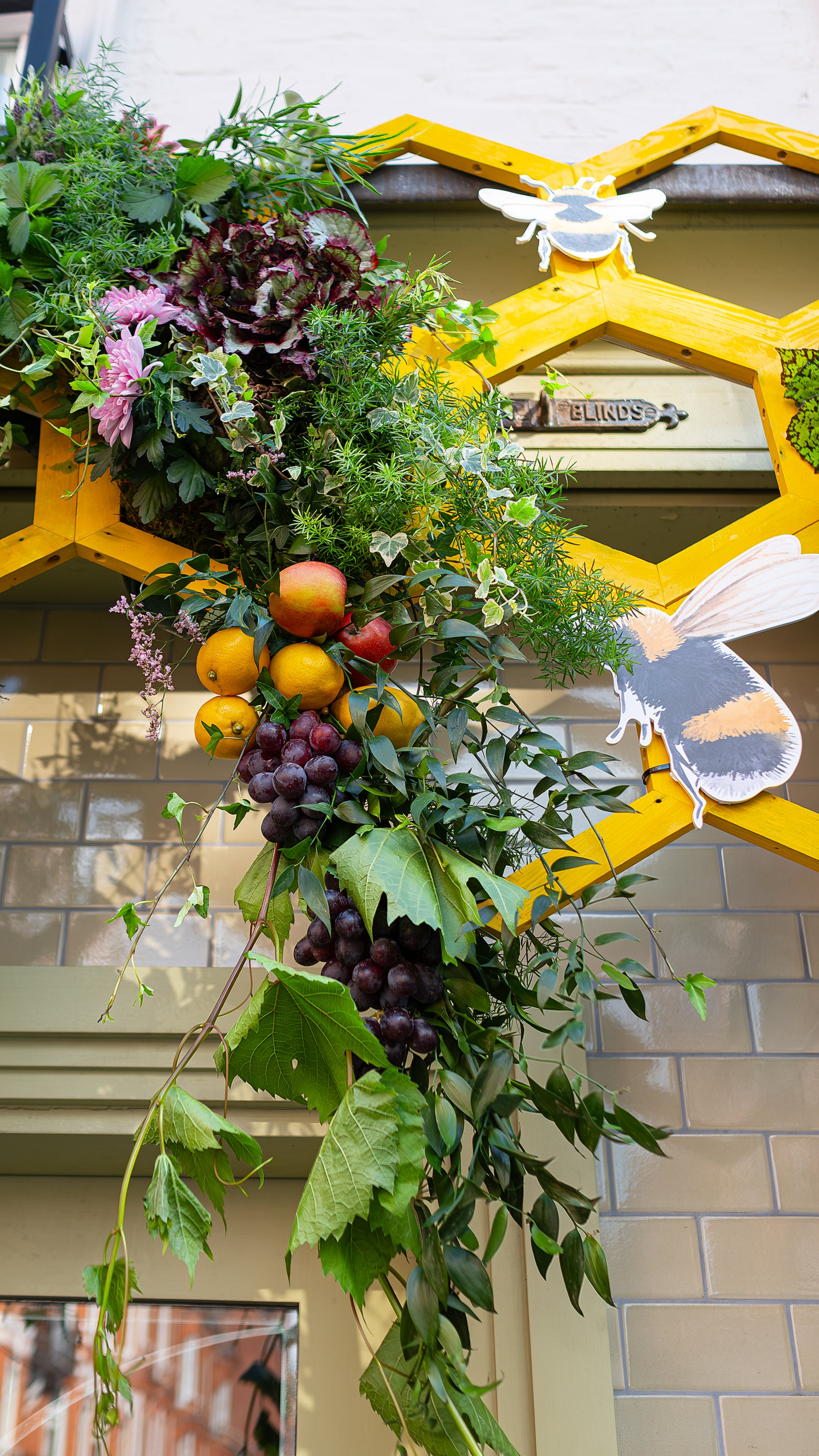 The Five Field Restaurant floral arrangement for Chelsea in Bloom features a vibrant mix of greenery, citrus fruits, and pink flowers artistically assembled around a yellow honeycomb structure, embodying a garden-to-plate freshness theme - designed, created and installed by event florist Amaranté London.