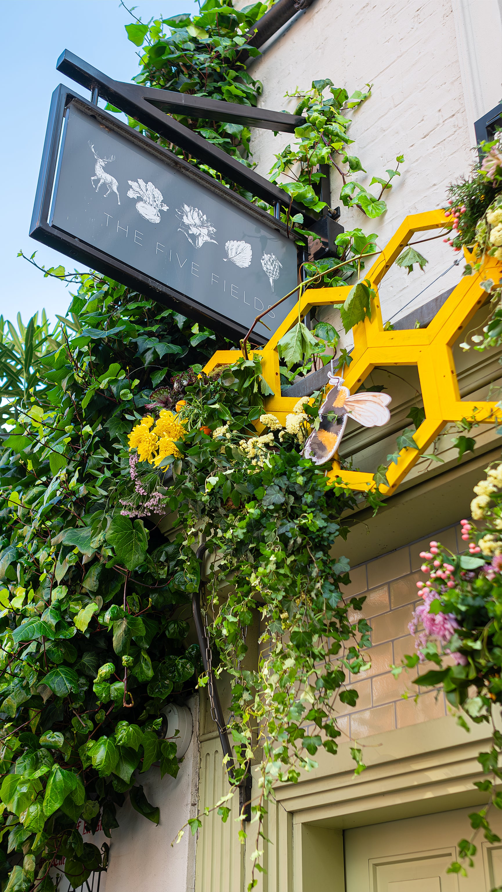 An elegant floral arrangement by Amaranté London at The Five Fields, with lush greenery, bright flowers, and oversized bee decorations against the restaurant sign, illustrates a nature-centric design for Chelsea in Bloom.