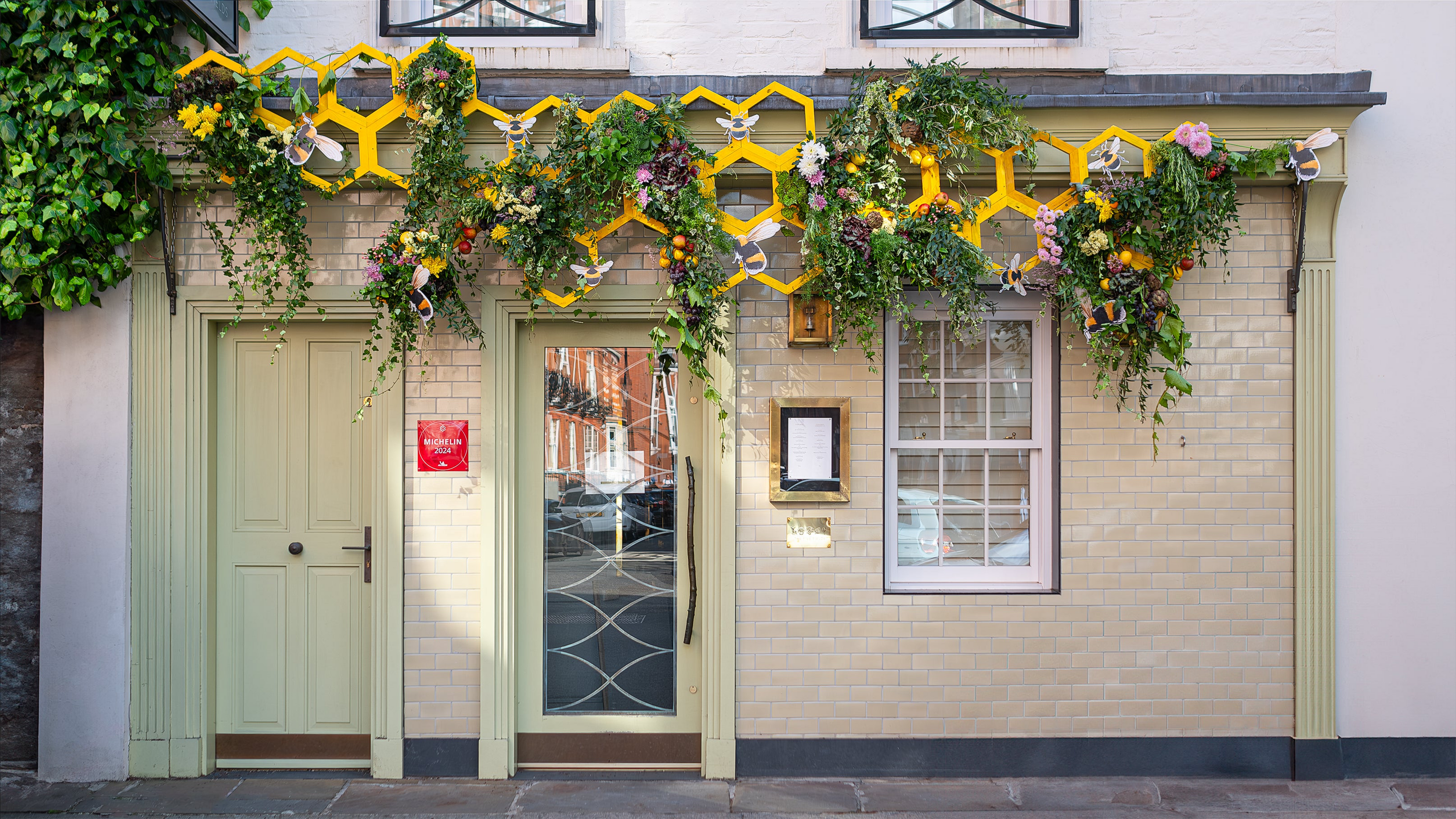 A bespoke floral installation at The Five Fields for Chelsea in Bloom by event florist Amaranté London showcasing an ornate storefront adorned with ivy, bright yellow geometric structures, and lush floral clusters interspersed with decorative bees.