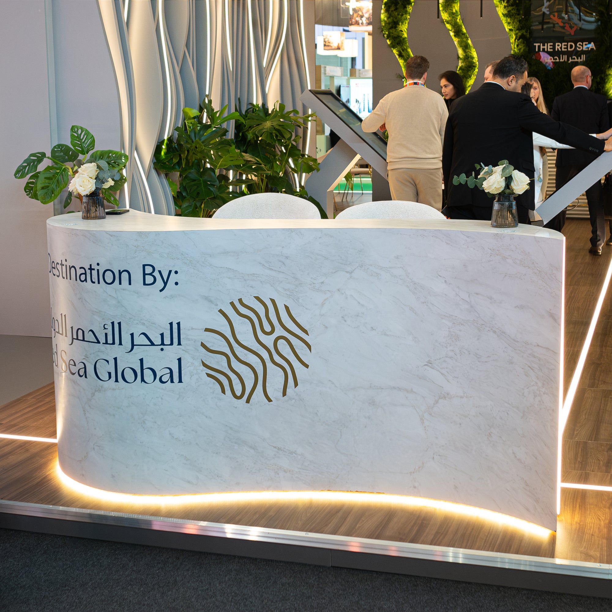 The sleek marble reception desk at WTM London for the Red Sea Global booth is adorned with a pure white rose arrangement in a stylish glass vase. In the background, a planter featuring large plants provides a professional and luxurious welcome for event guests.