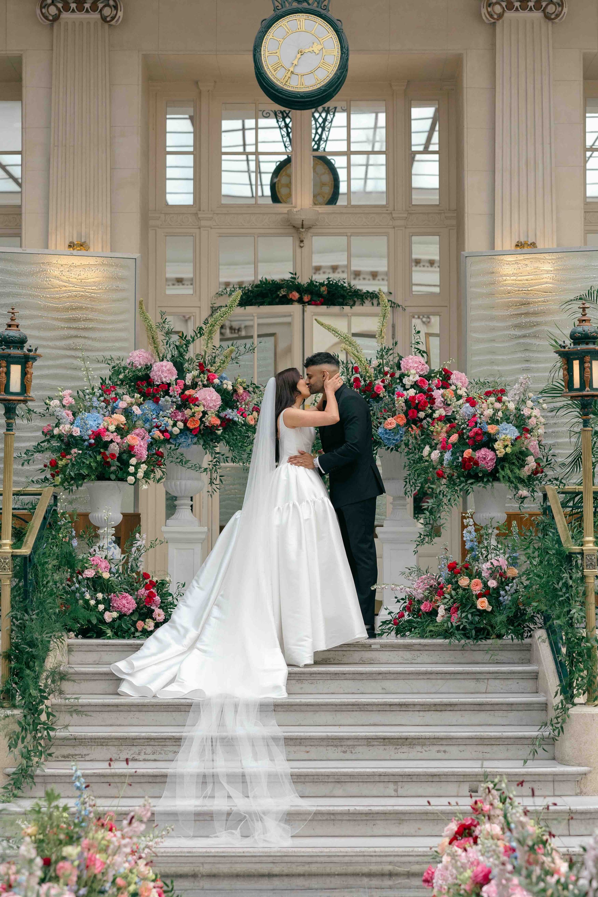 Wedding ceremony kiss on grand staircase with luxury floral installations by Amaranté London production house