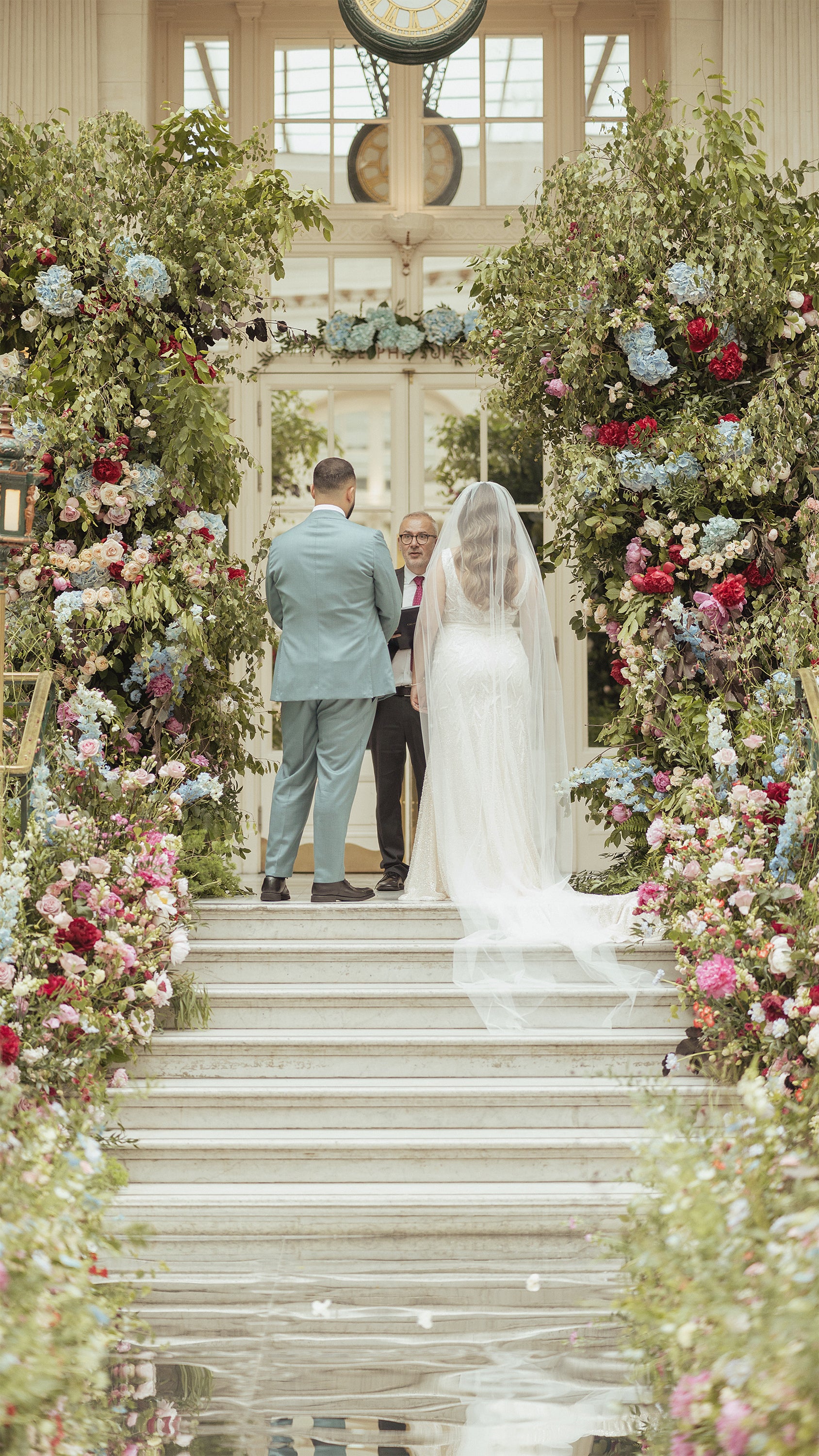 ALT: "Wedding ceremony at Waldorf Hilton with bride, groom, and officiant under elaborate floral arch on marble steps with classical architecture - Floral Design by Amaranté London