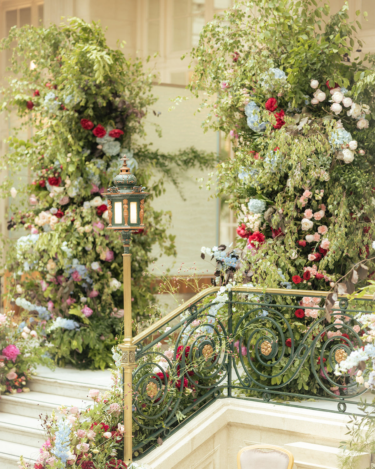 Romantic staircase floral installation for a luxury wedding venue, featuring red, white, and blue blooms with trailing greenery adorning ornate balustrade and lantern details - Amaranté London, luxury wedding event floristry