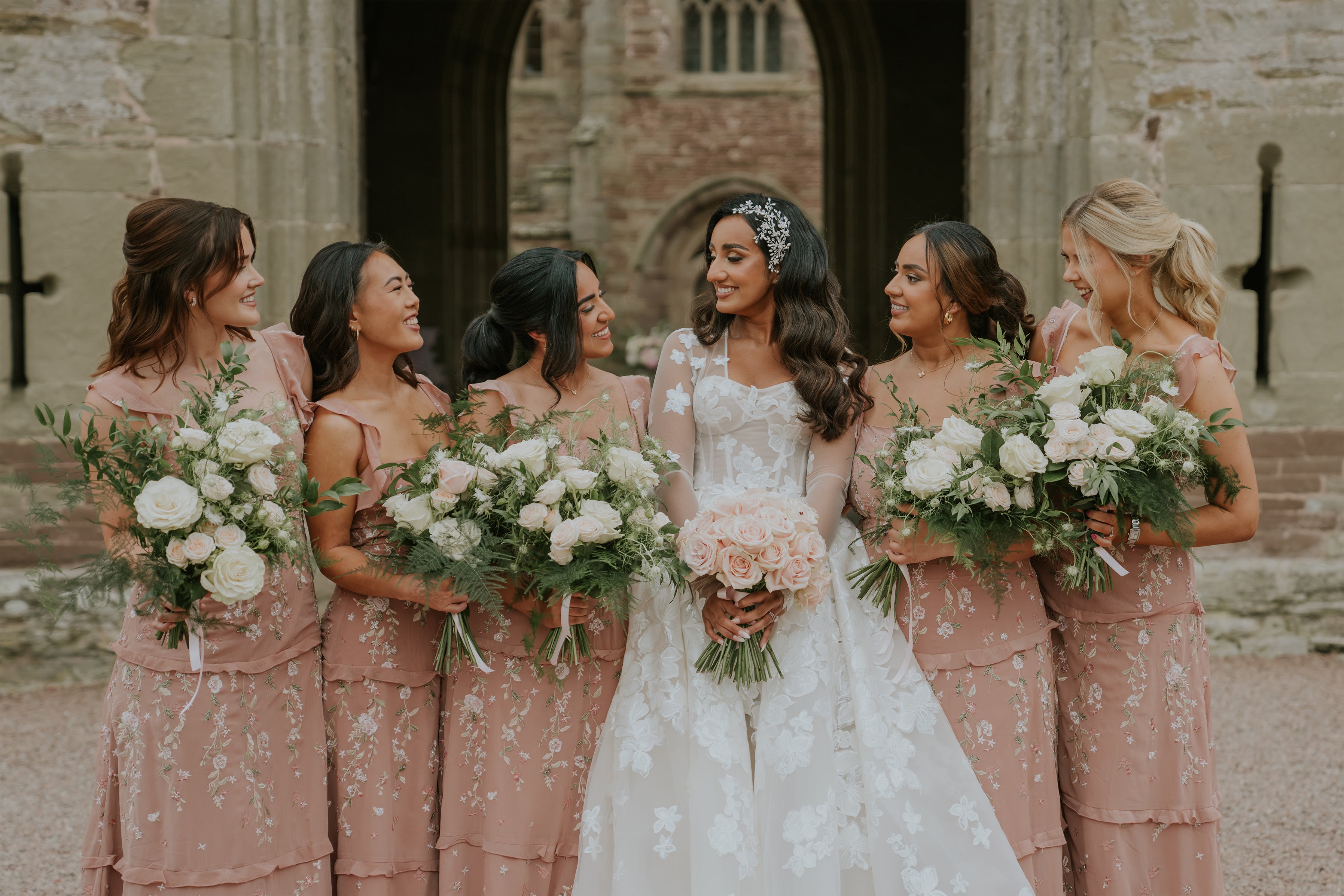 Coordinated bridal party bouquets featuring white roses and blush accents with natural greenery against a historic stone abbey backdrop - Amaranté London, floral design for luxury wedding floristry