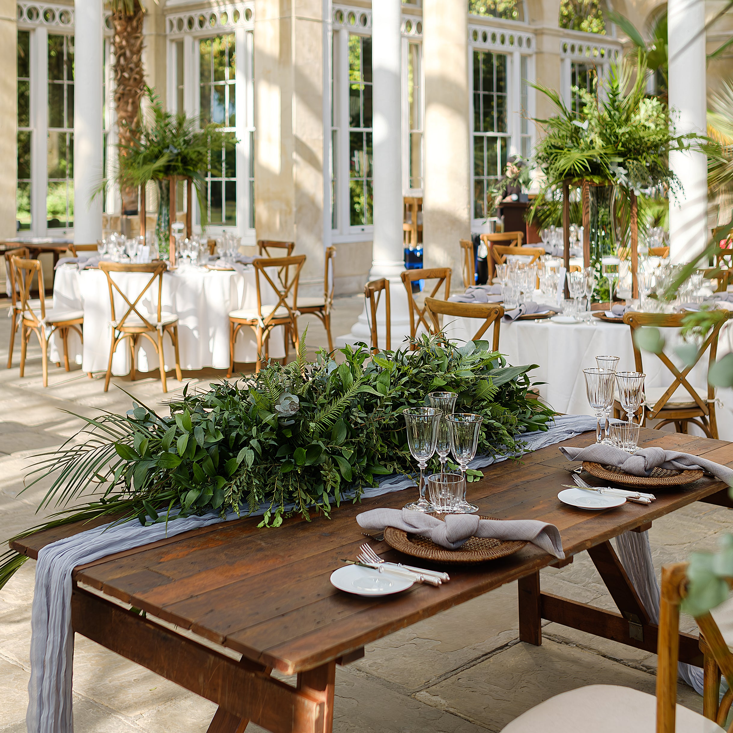 Natural greenery table runner with mixed foliage and ferns creating an organic centerpiece on a rustic wooden table in a bright wedding setting - Amaranté London