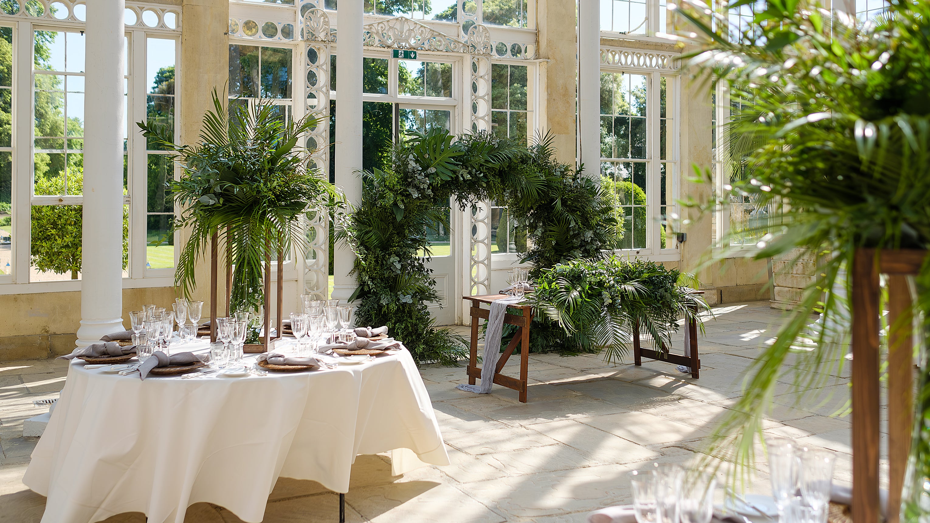 Tropical foliage as a backdrop with palm fronds and mixed greenery creating a natural archway in a bright wedding venue - Amaranté London