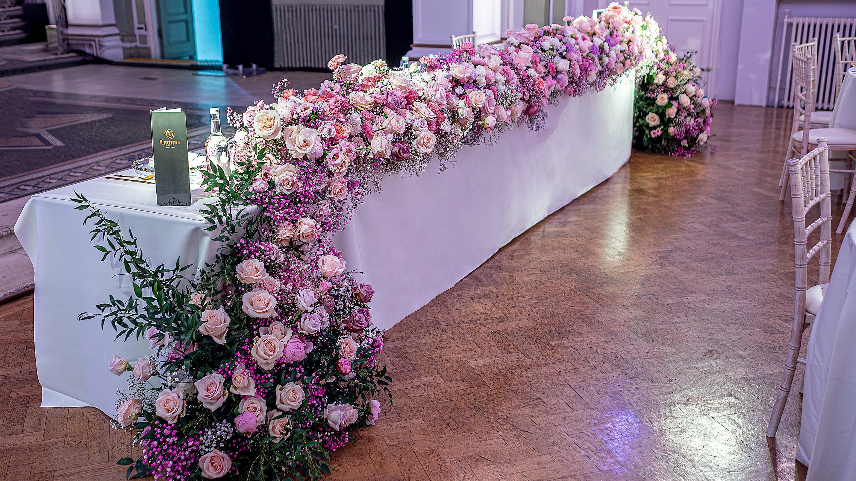 Continuous floral table runner featuring blush roses and purple waxflower, creating seamless design along white-draped tables in grand wedding venue - Wedding event styling and elegant tablescape arrangements by Amaranté London