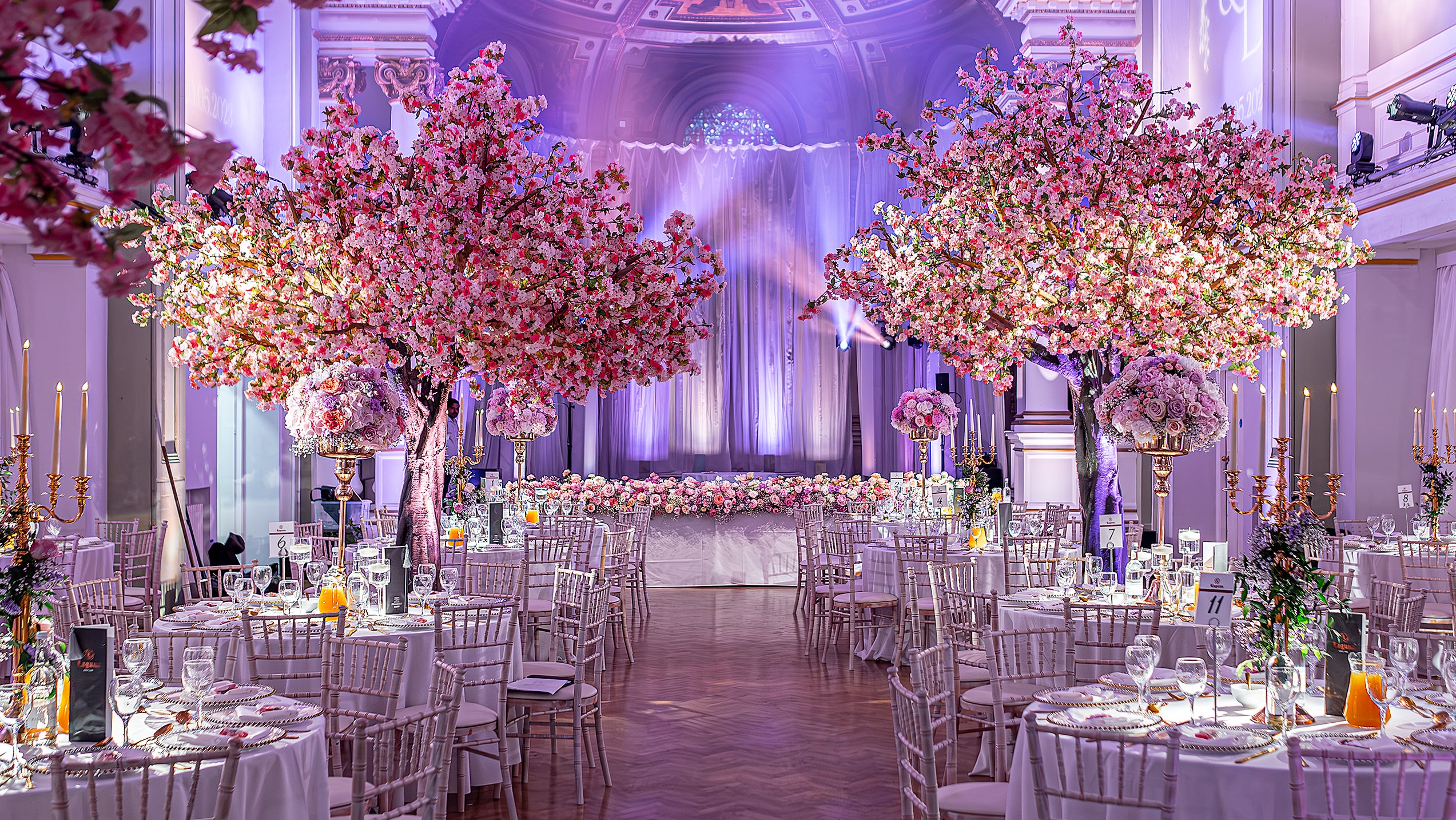 Spectacular ballroom transformation of a wedding venue featuring towering cherry blossom trees and coordinated pink floral centerpieces with elegant table settings - Amaranté London
