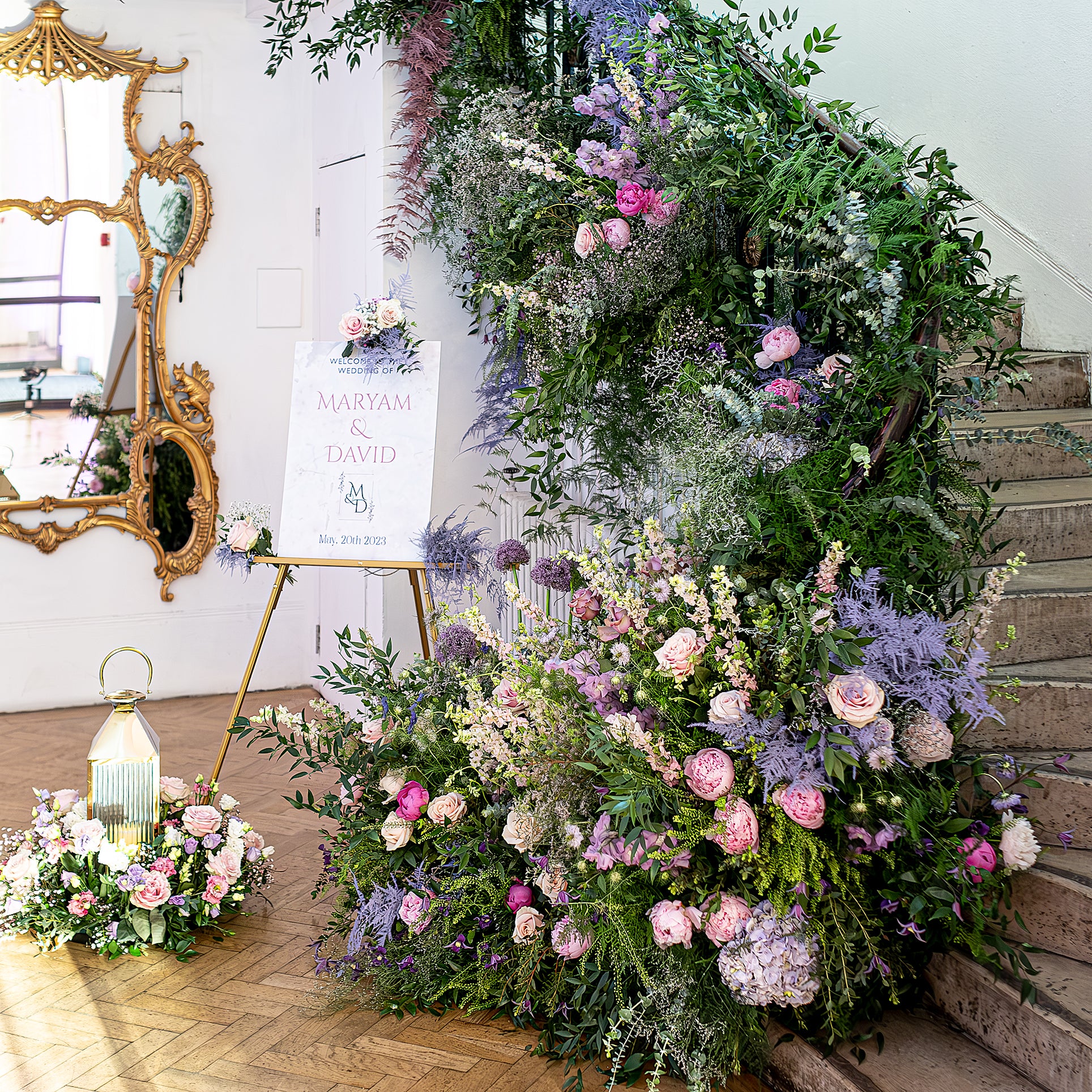  Staircase floral installation for a wedding featuring purple and pink blooms with mixed greenery cascading down steps alongside welcome signage - Floral Design by Amaranté London