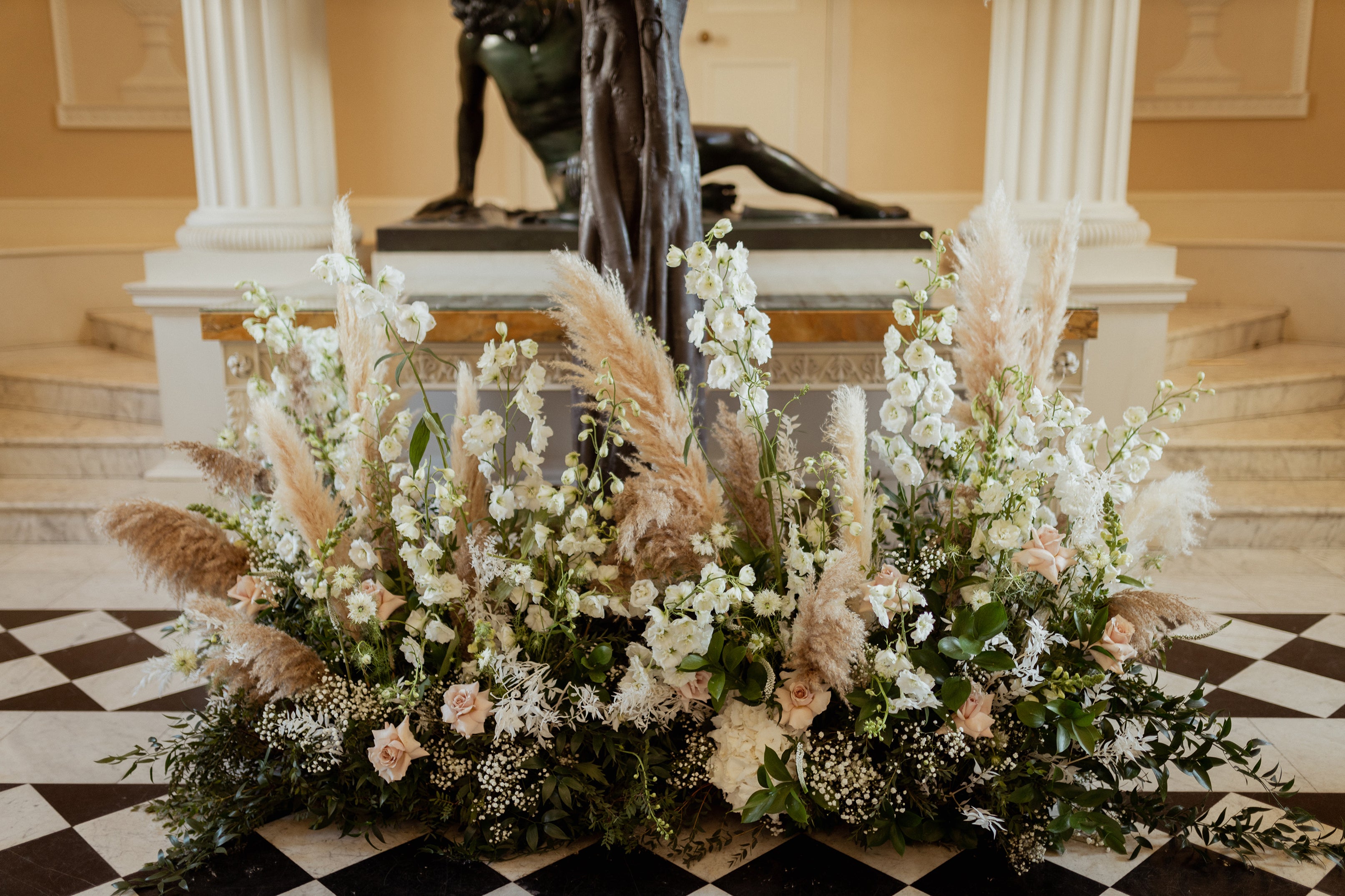 Ground-level floral arrangement with white delphiniums, blush roses, and pampas grass, creating a base installation beneath a classical statue - Amaranté London, Floral Design for luxury wedding venues