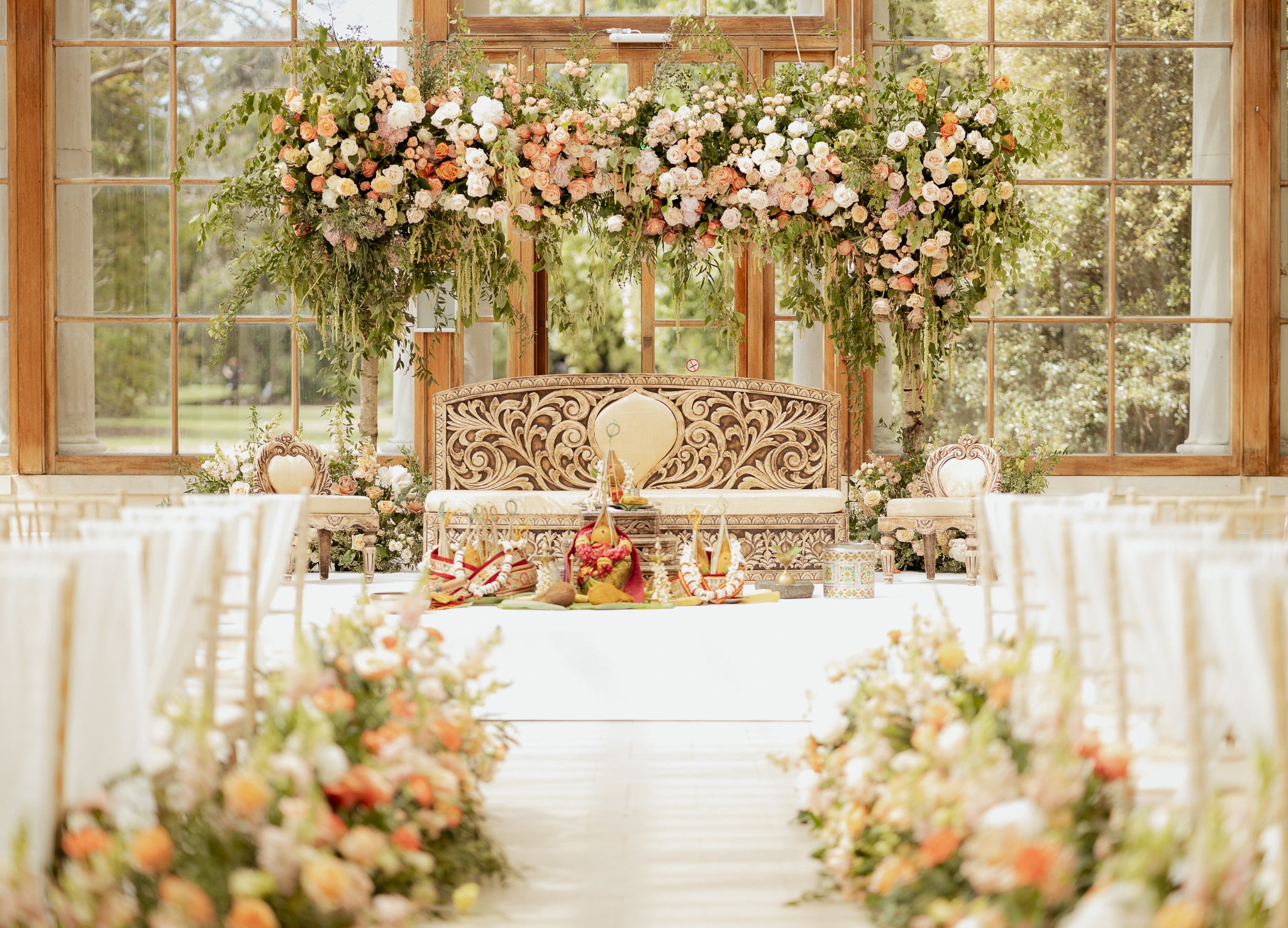 Luxurious ceremony backdrop featuring cascading floral installation with peach roses, white blooms, and trailing greenery above an ornate seating area, demonstrating Amaranté London's grand floral archways that frame weddings