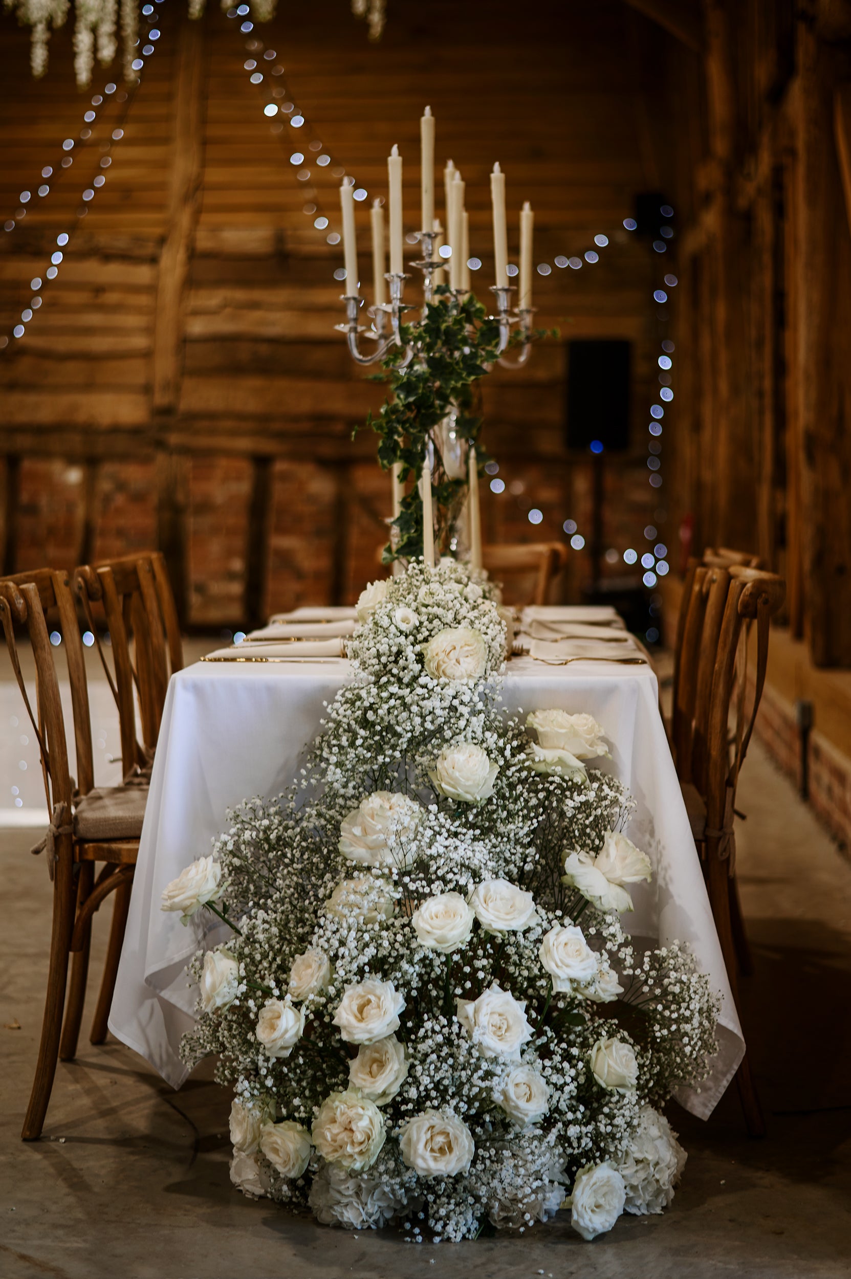 Romantic table runner featuring white roses and baby's breath, creating a flowing centerpiece with a tall candelabra in a rustic barn setting - Amaranté London, elegant tablescape design for intimate wedding atmospheres