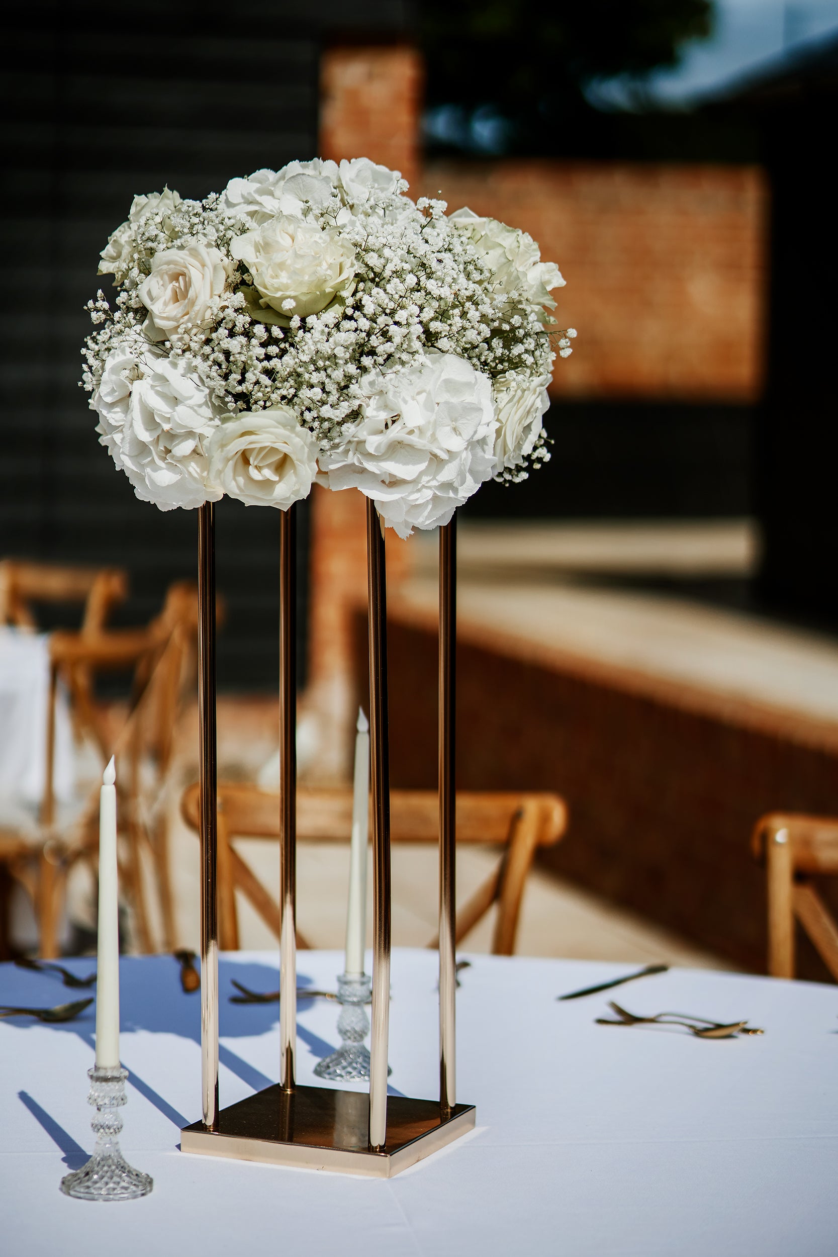 Elevated white rose and baby's breath arrangement on a modern geometric stand, creating sophisticated height variation in a rustic barn venue - Amaranté London, contemporary floral design for weddings