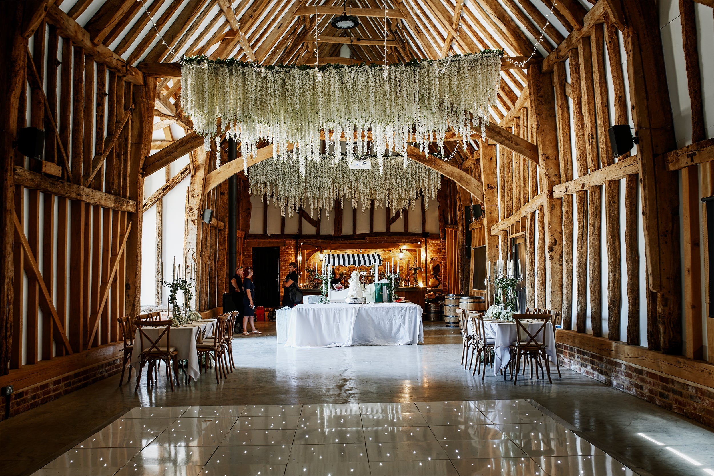 Overhead floral installation with cascading white blooms and greenery suspended above rustic barn venue dining area - Amaranté London