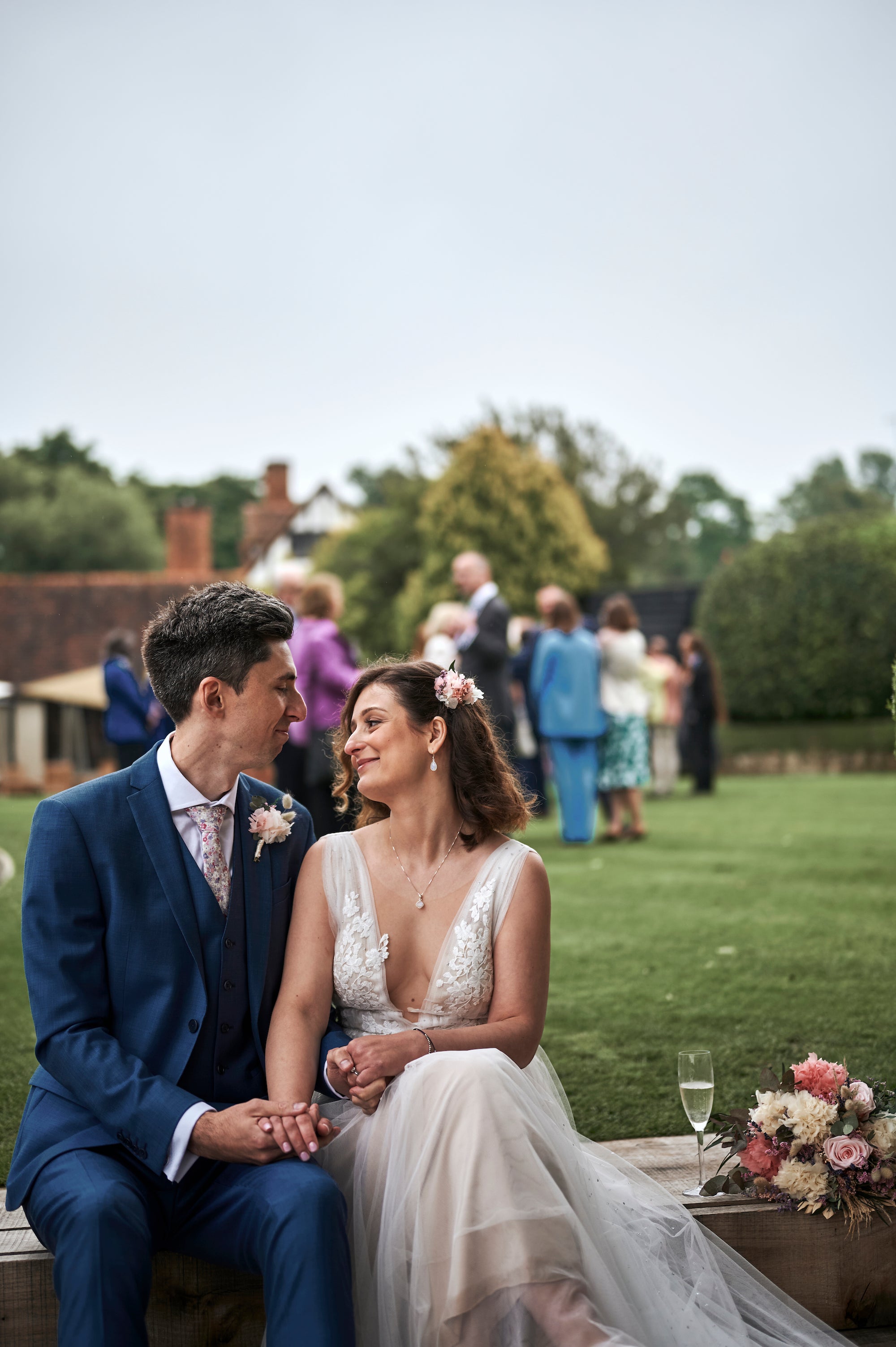 Intimate couple moment with coordinated coral boutonniere and hair flower details in a natural garden reception setting - Amaranté London, floral arrangements for luxury weddings