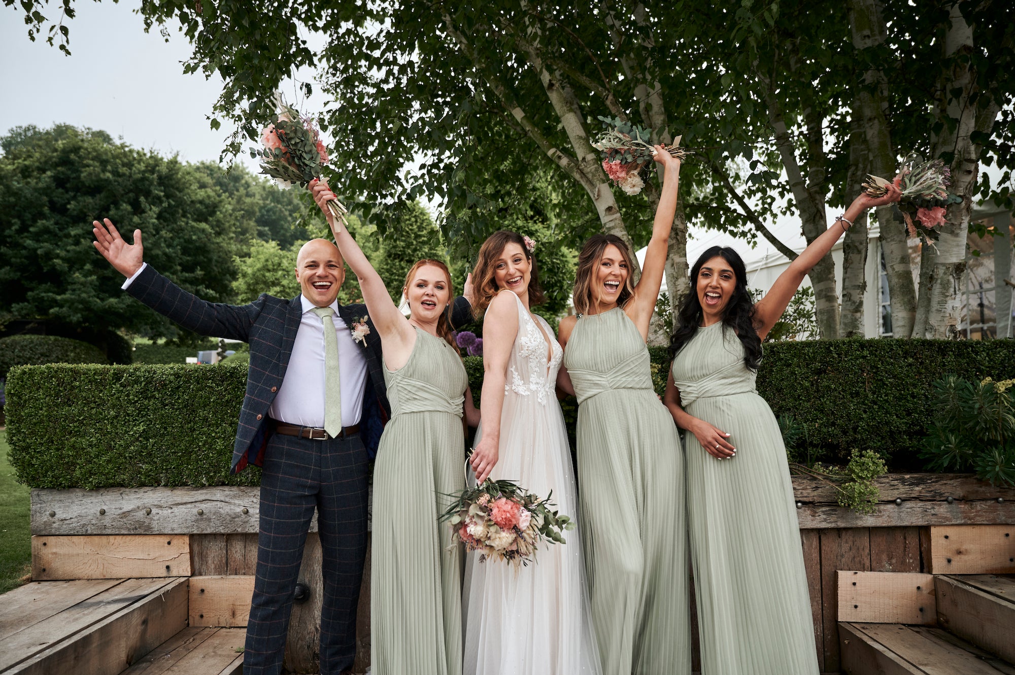 Celebratory bridal party with coordinated coral and peach bouquets featuring roses and greenery in a natural garden setting - Amaranté London