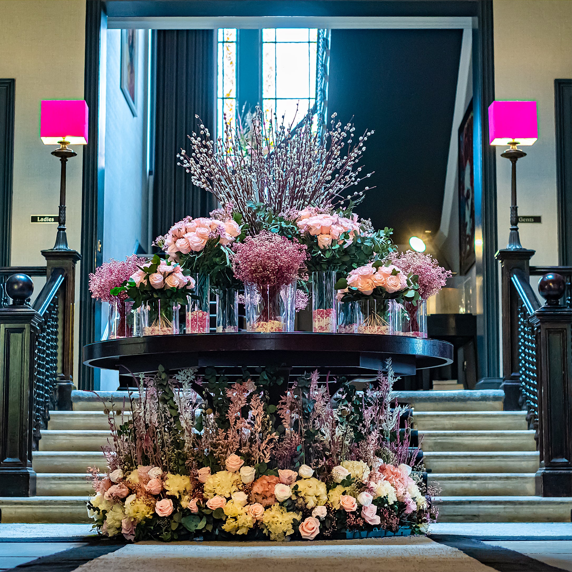 Multi-tiered foyer floral display featuring peach roses, pussy willow branches, and seasonal blooms arranged on a round table with base installation - Amaranté London, Floral Design for luxury wedding venues