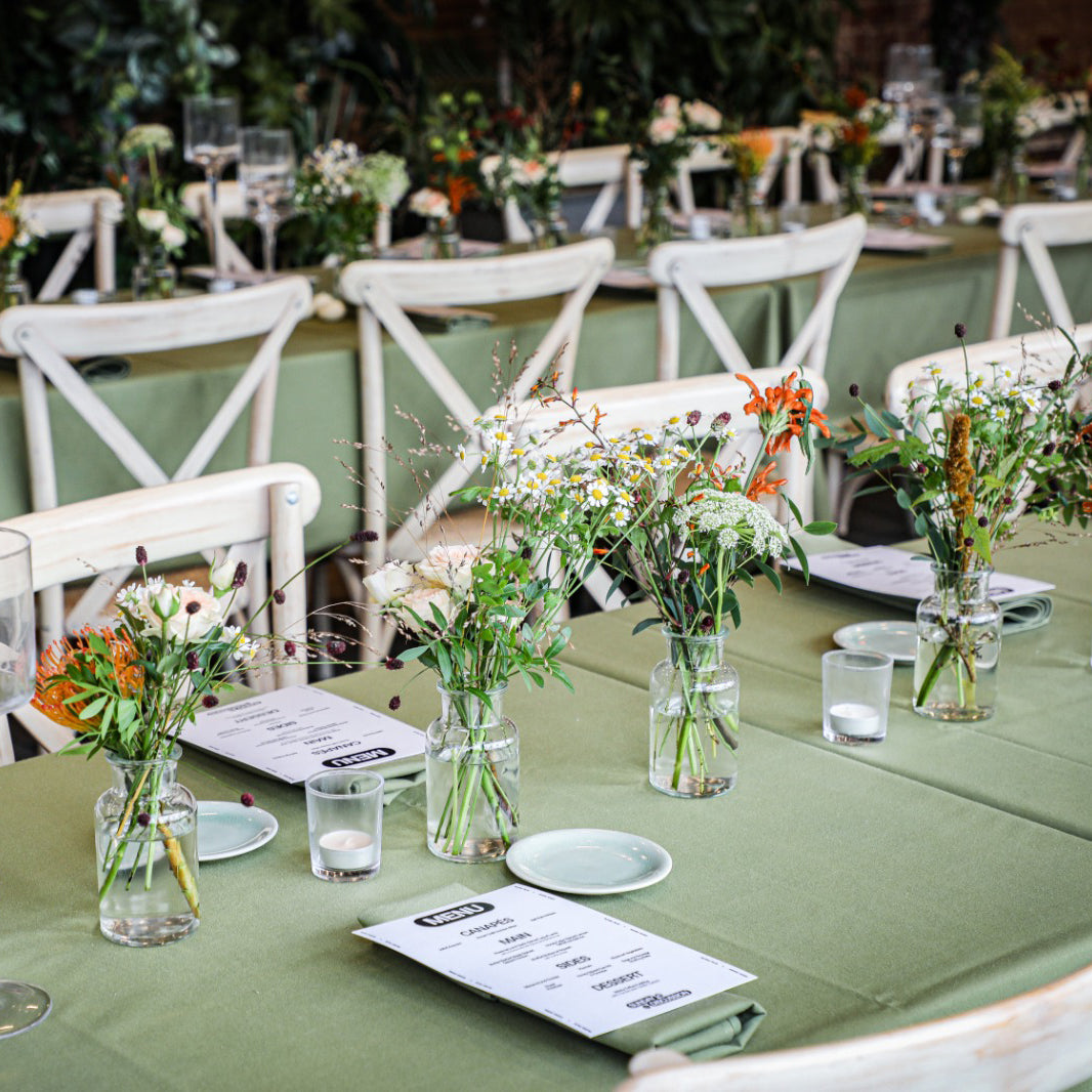 Natural wildflower-style table arrangements for a wedding featuring mixed seasonal blooms and grasses in simple glass vases on sage green linens - Floral Design by Amaranté London Event Florist