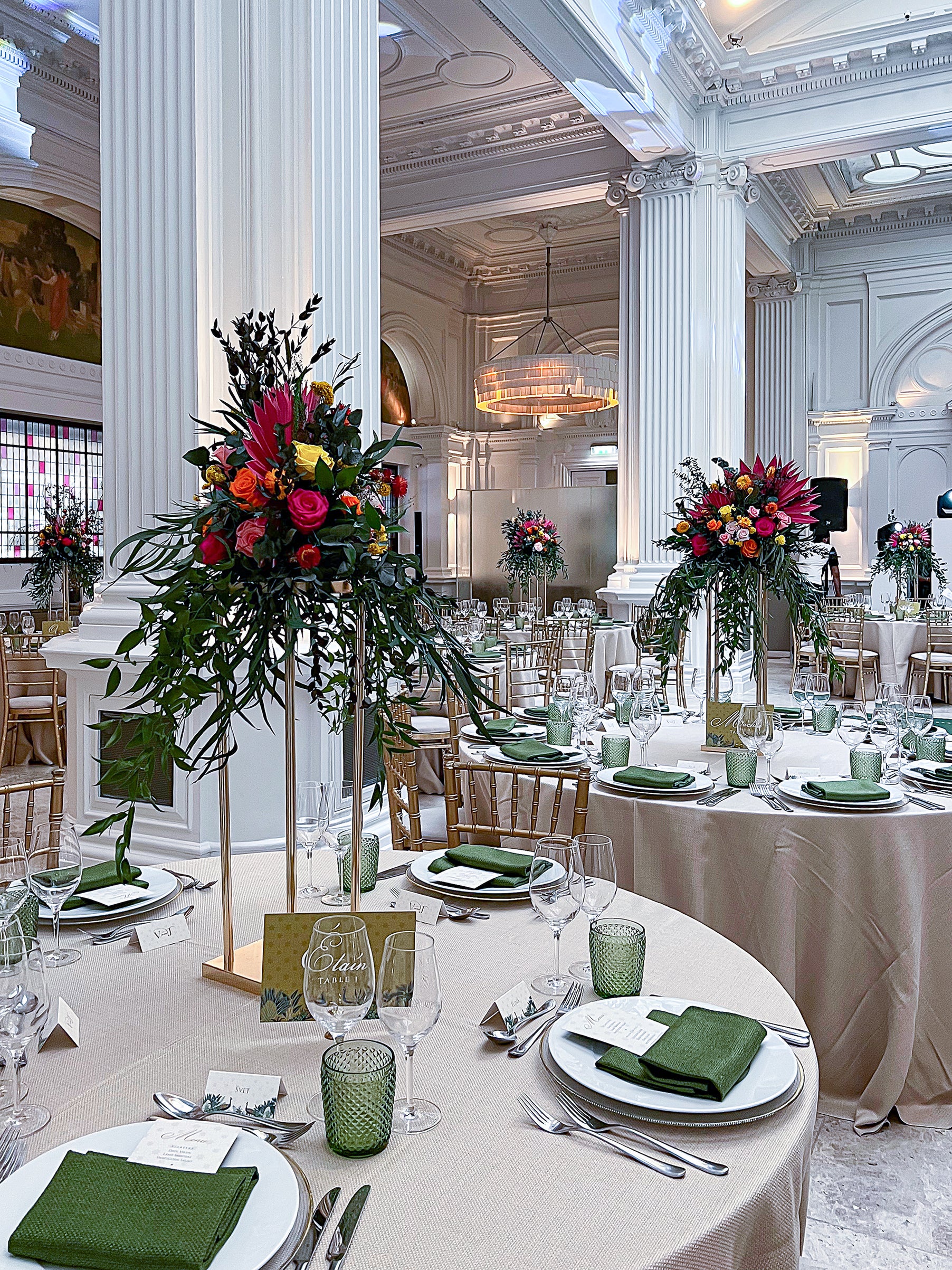 Elevated bespoke floral centerpieces for a wedding event, featuring vibrant pink roses and dark foliage on tall gold stands in a grand neoclassical dining hall with coordinated green table settings - Amaranté London Event Florist