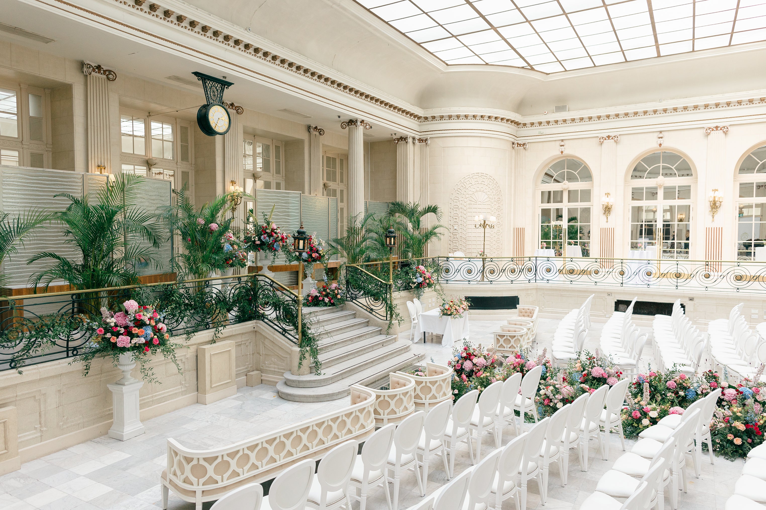 Grand ceremony space featuring tropical palm arrangements and colorful floral installations on a marble staircase with formal seating in a neoclassical luxury wedding venue - Floral Design by Amaranté London