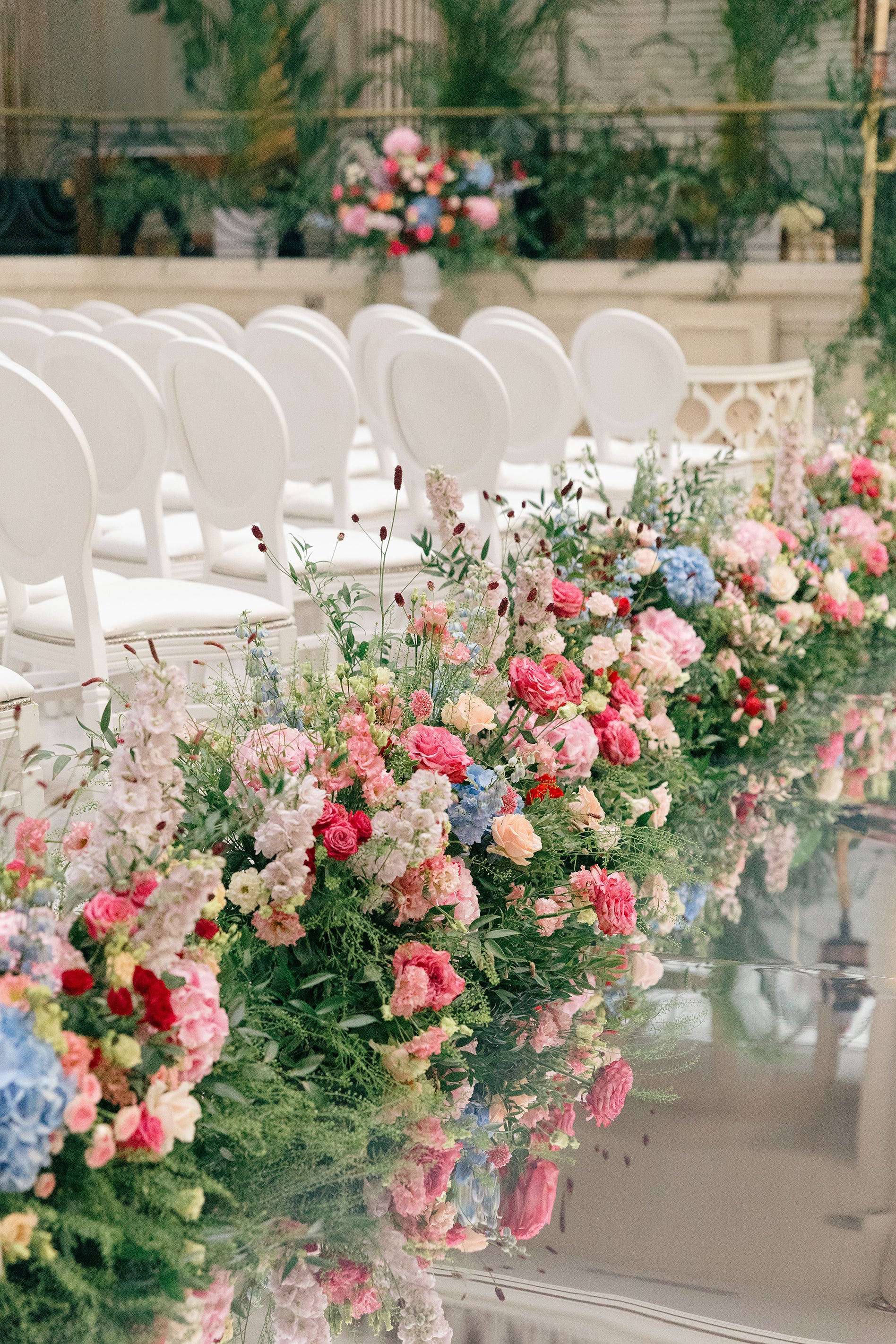 Abundant mixed bespoke floral arrangements with pink roses, blue hydrangeas, and wildflowers create a romantic aisle border on a reflective marble surface in a classical wedding venue, creating a lush garden-style look - Floral Design by Amaranté London