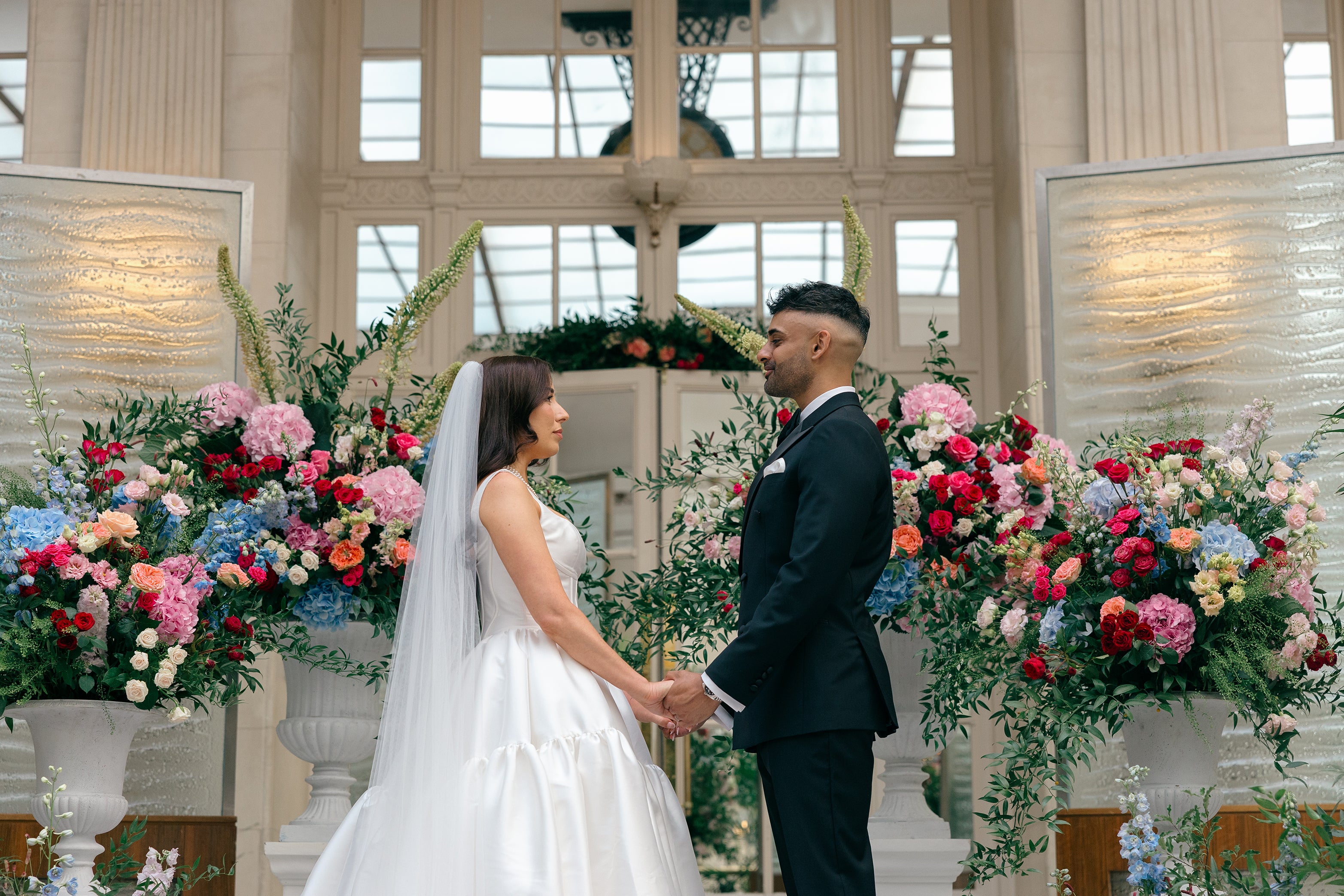 Two large floral displays featuring vibrant mixed blooms, including pink hydrangeas, red roses, and blue accents, positioned on classical pedestals - Floral Design by Amaranté London