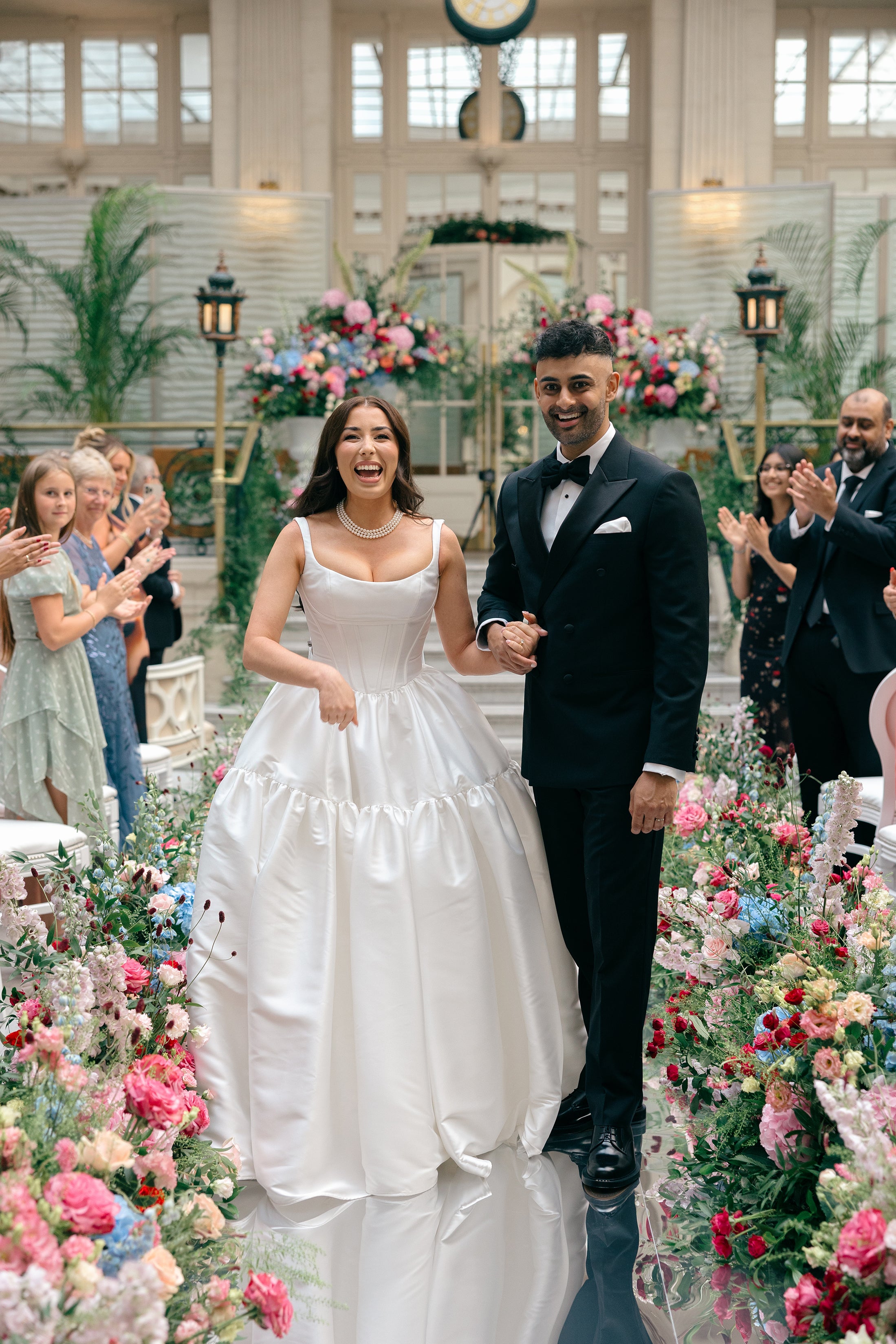 Vibrant aisle floral arrangements featuring pink, blue, and coral blooms creating a colourful pathway in an elegant glass-roofed wedding venue - Floral Design by Amaranté London