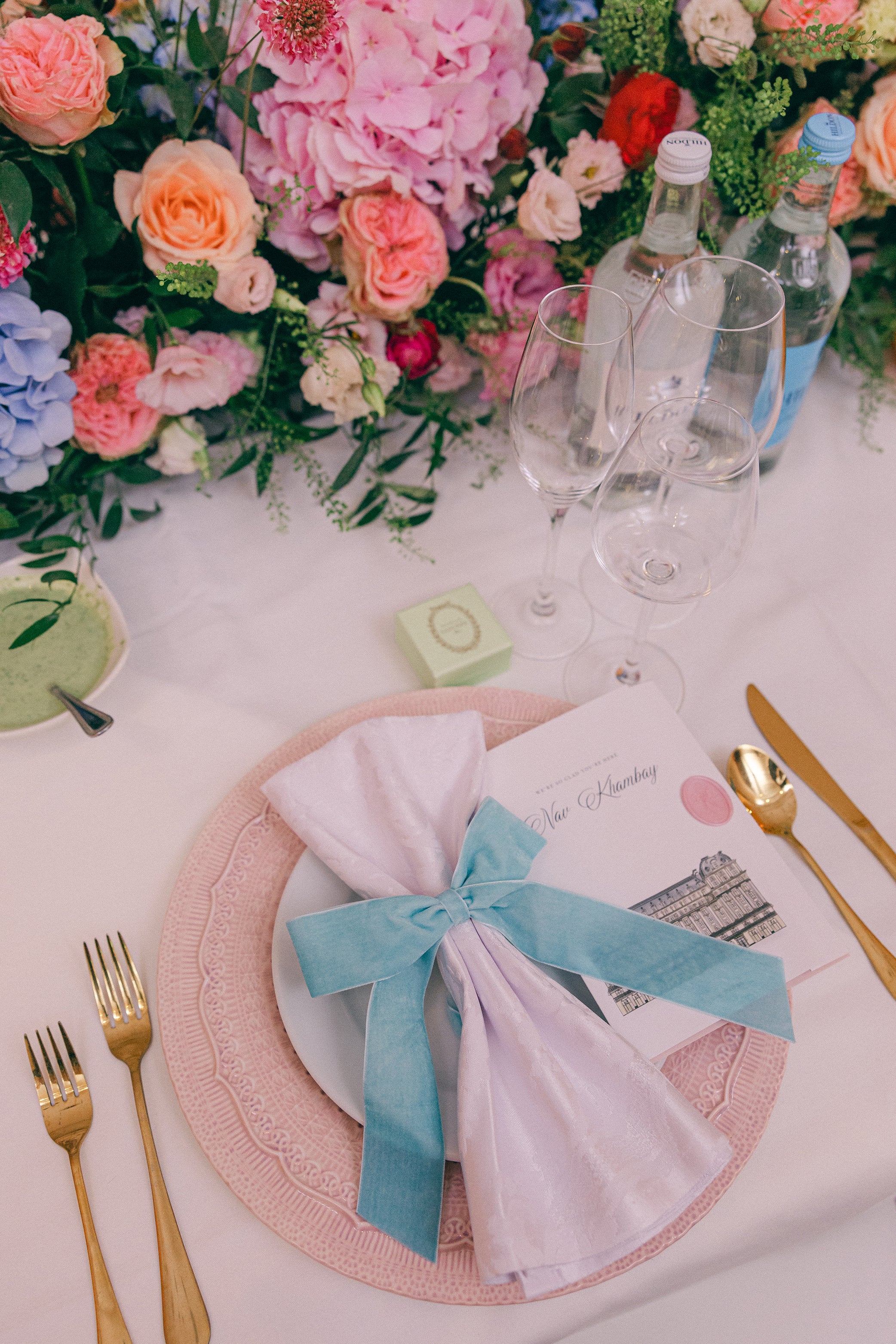 Detailed place setting with floral centerpiece featuring pink peonies, coral roses, and textural elements alongside elegant pink glassware and blue ribbon accents - Floral Design by Amaranté London