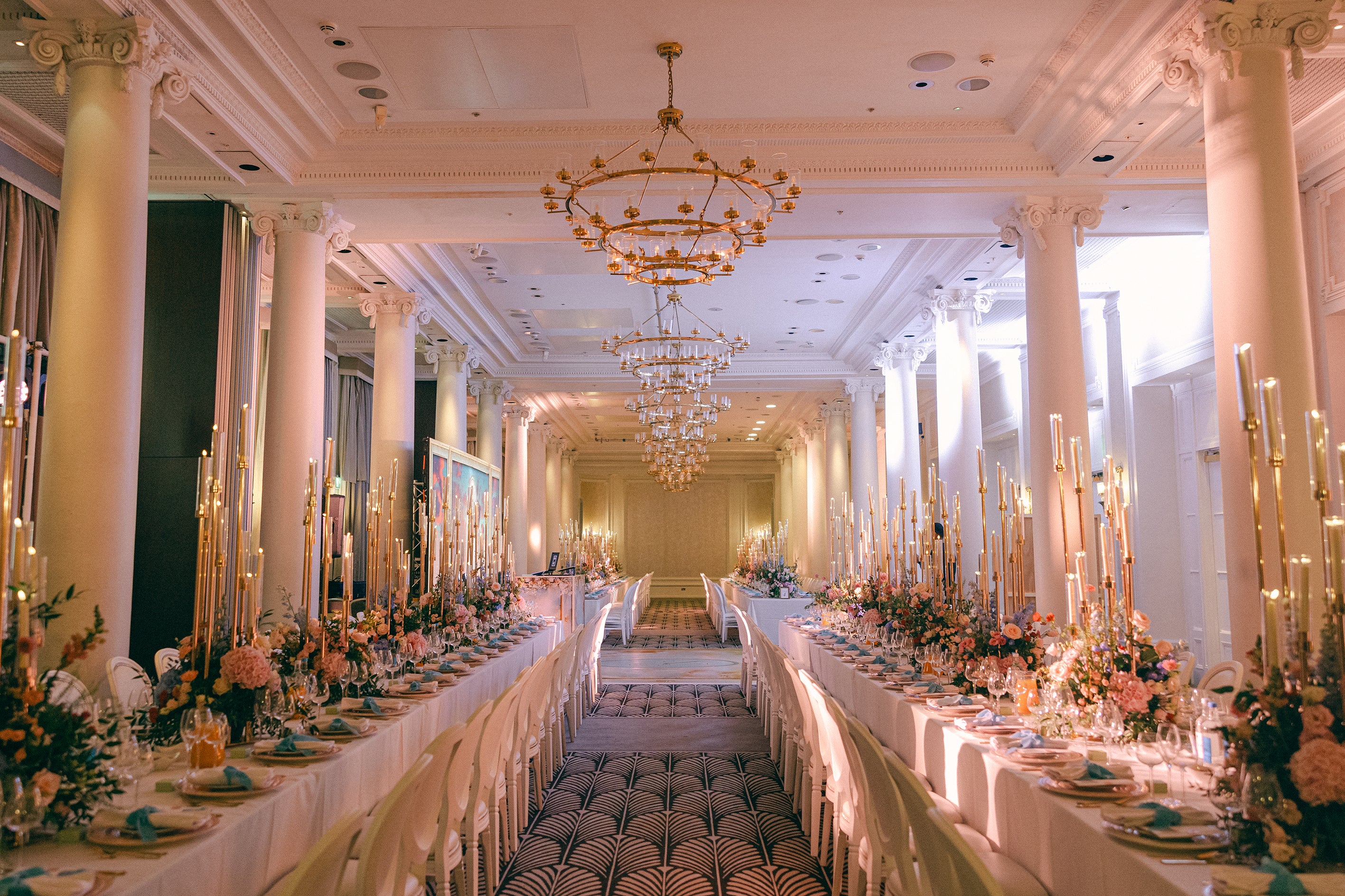 Spectacular ballroom dining setup with towering candle installations and coordinated bespoke floral centerpieces creating dramatic golden lighting throughout the grand columned wedding venue - Floral Design by Amaranté London
