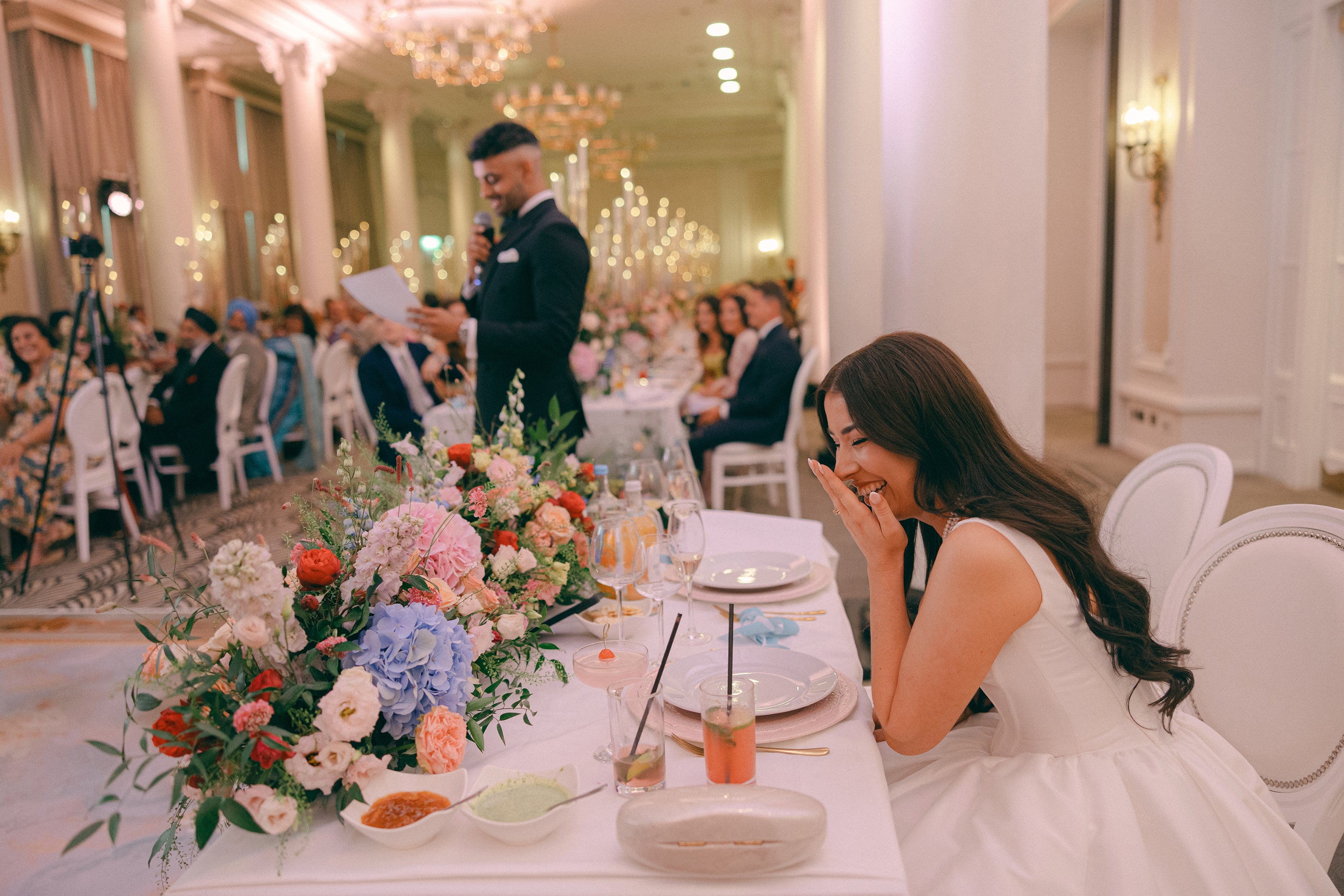 Joyful reception moment, a warm atmosphere in an elegant ballroom setting decorated with wedding flowers: colorful bespoke floral centerpieces featuring pink, coral, and blue blooms - Floral Design by Amaranté London