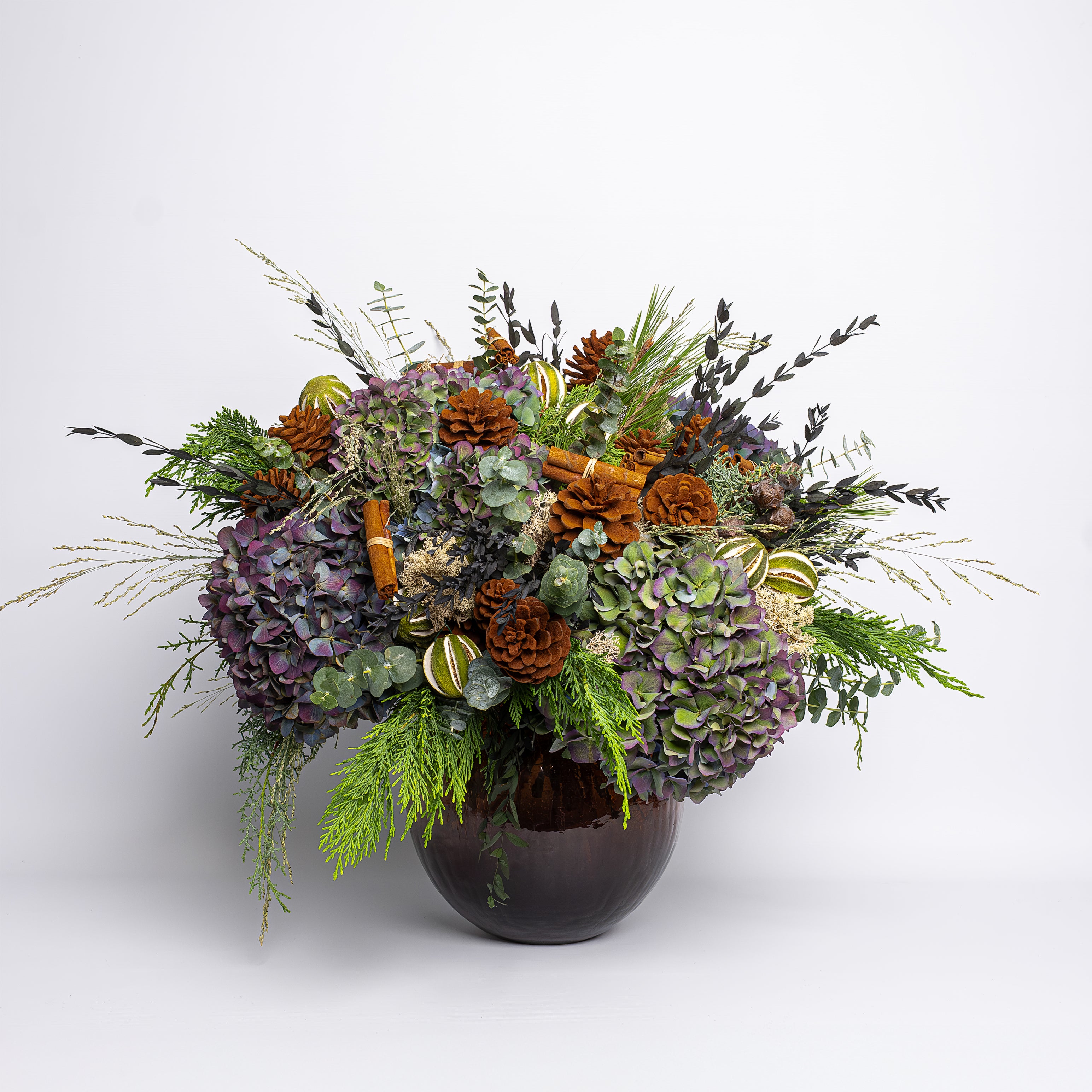 Decorative floral arrangement with greenery and pinecones in a brown vase on a light gray background