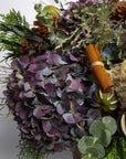 Decorative wreath with purple flowers, greenery, and cinnamon sticks on a white background