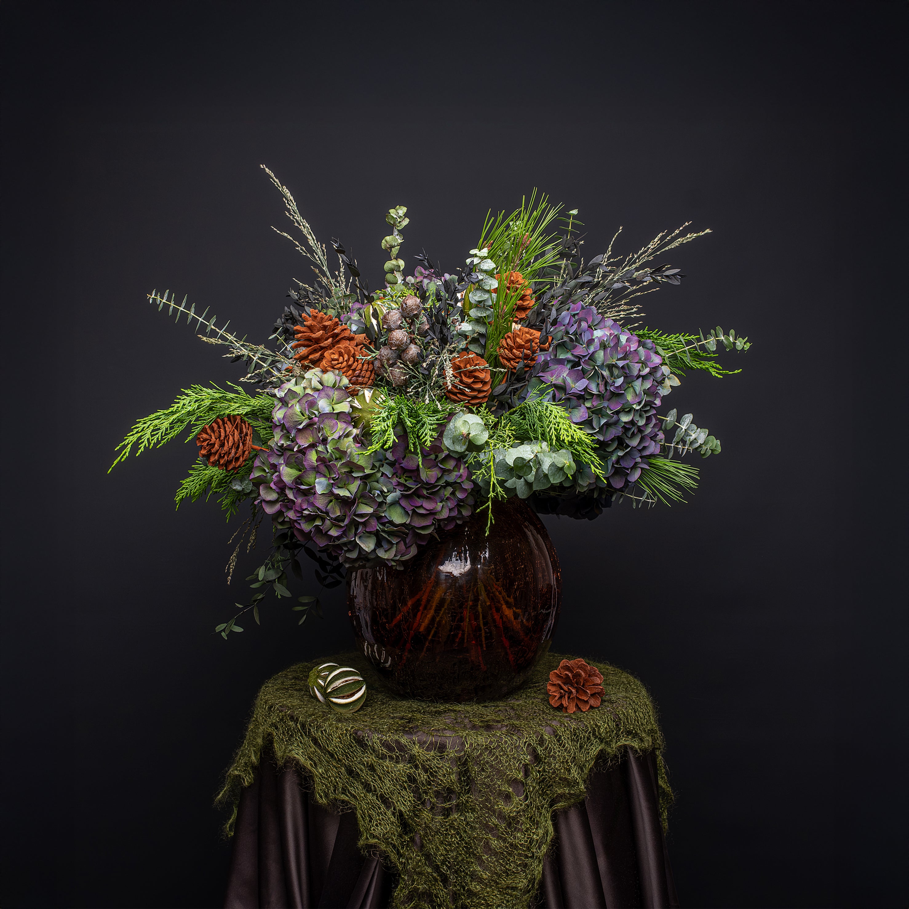 Modern Christmas flower arrangement featuring purple and mauve hydrangeas, rust-colored dried floral stems, fresh evergreen pine, ornamental dried grasses, and silver eucalyptus foliage arranged in a dark glass vase on a moss-draped surface with dark fabric and festive ornamental accents - Amaranté London