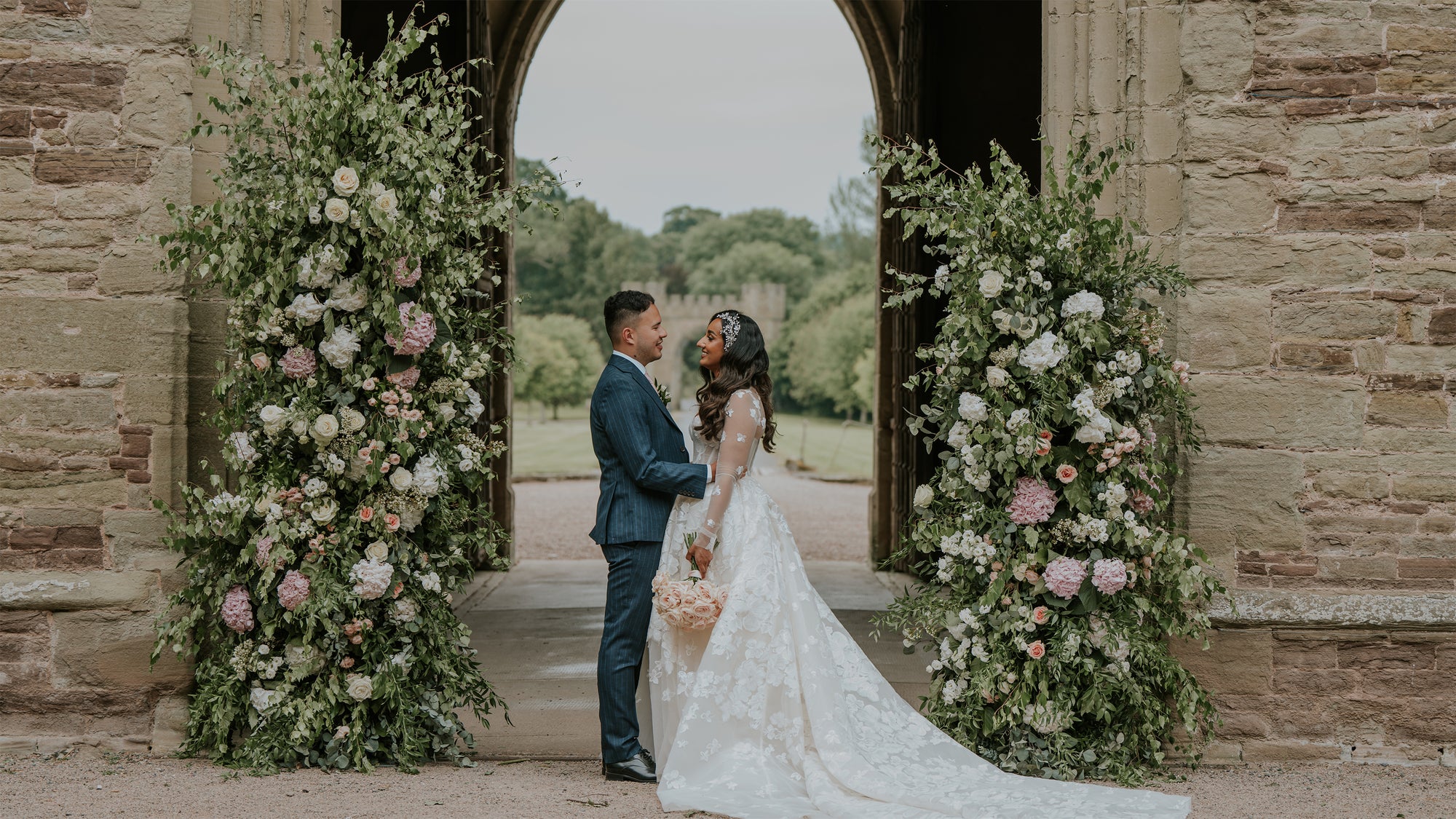Bride and groom embracing under ornate floral arch with white roses, pink hydrangeas and eucalyptus greenery framing Hampton Court Castle's historic stone archway - Floral Design by Amaranté London.