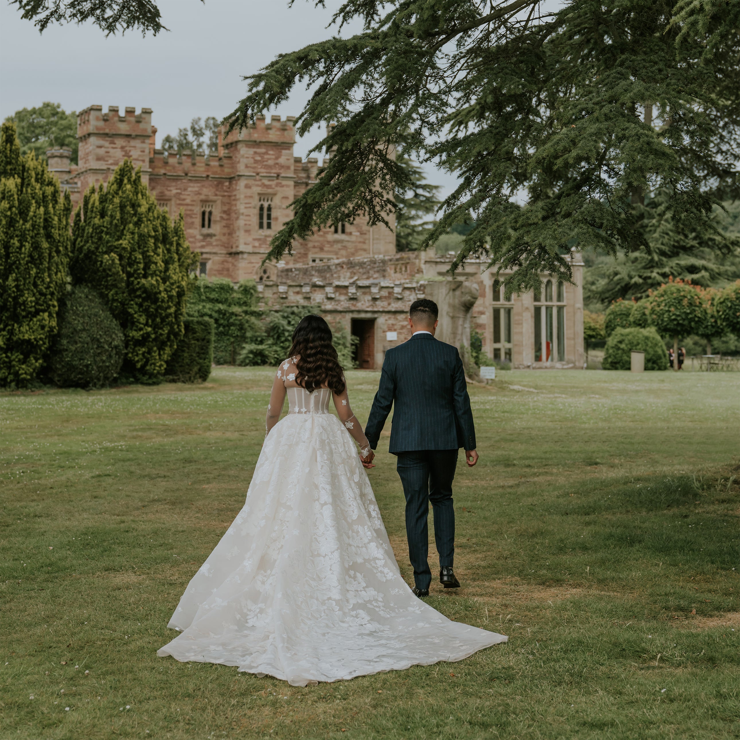 Bride and groom walking hand-in-hand across Hampton Court Castle's manicured lawns with the historic 15th-century stone castle and towers in background