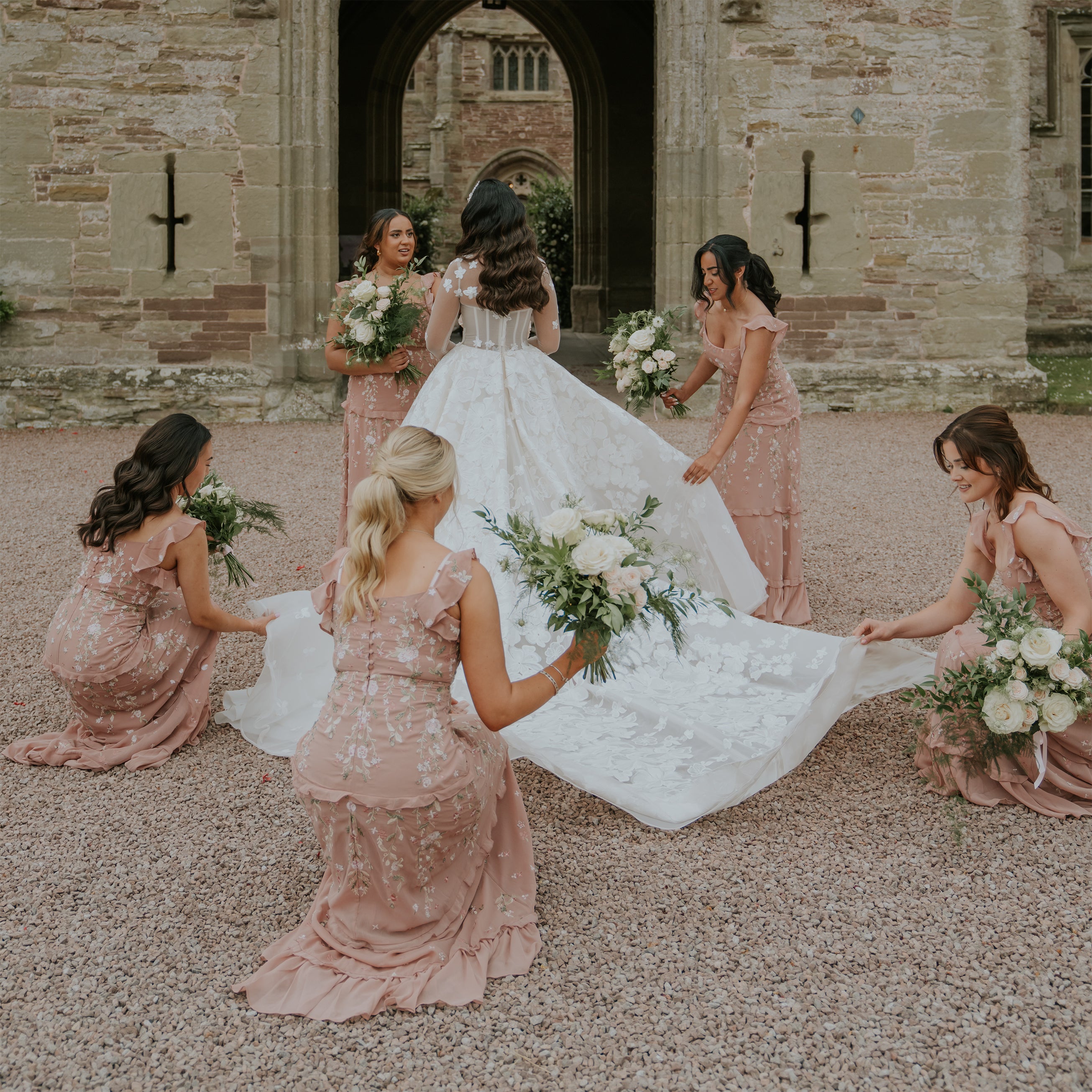 Bridesmaids in blush pink dresses holding white rose and eucalyptus bouquets while arranging the bride's train at Hampton Court Castle's stone courtyard - Floral Design by Amaranté London.